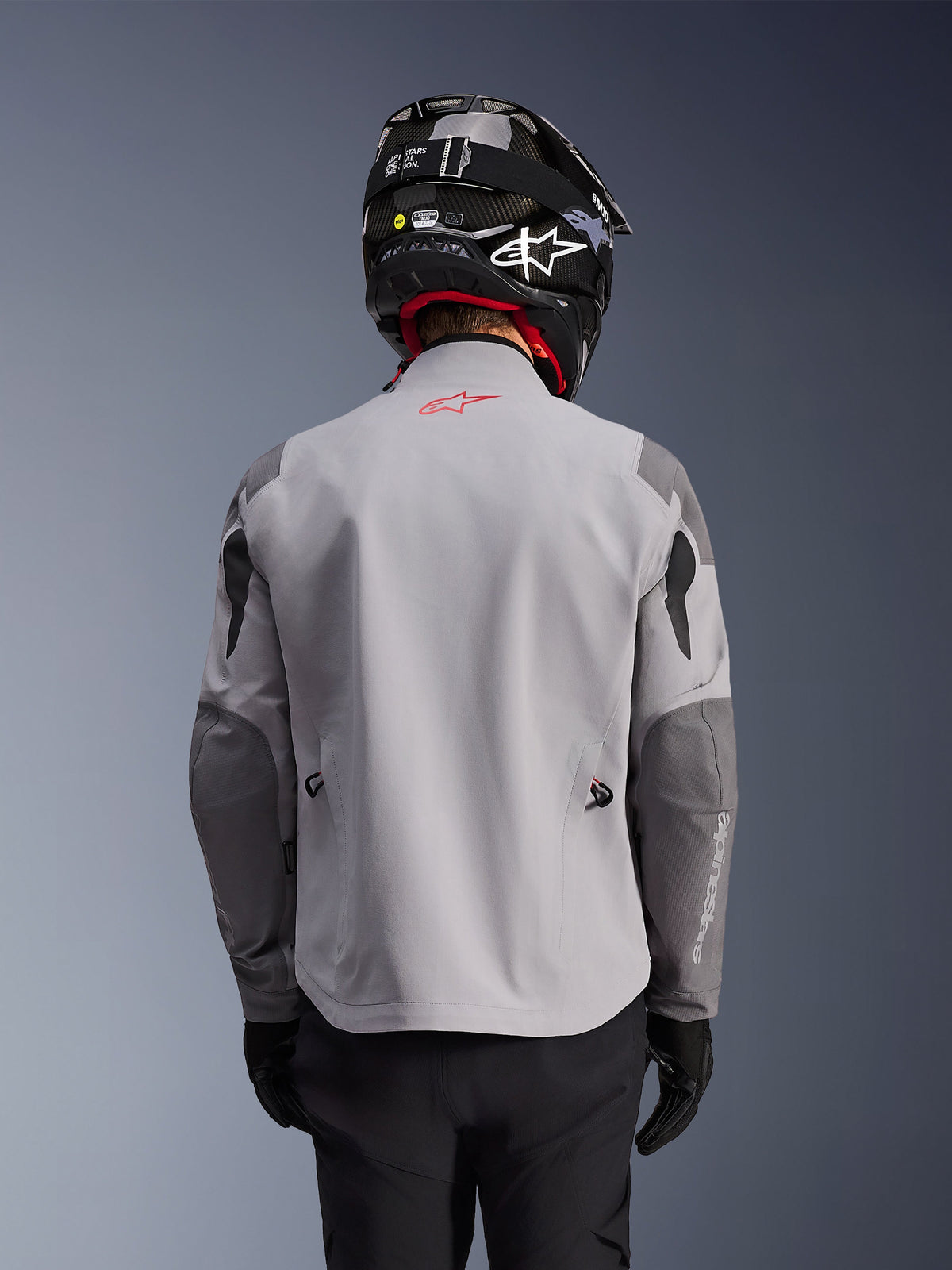 Alpinestars Maxdura Drystar®XF Laminated Armored Shell, Adventure Motorcycle Jacket, Mid Gray and Dark Gray, back view showing light gray body with darker gray reinforced panels on shoulders and elbows, red Alpinestars logo on the upper back, worn with a black helmet and black gloves.