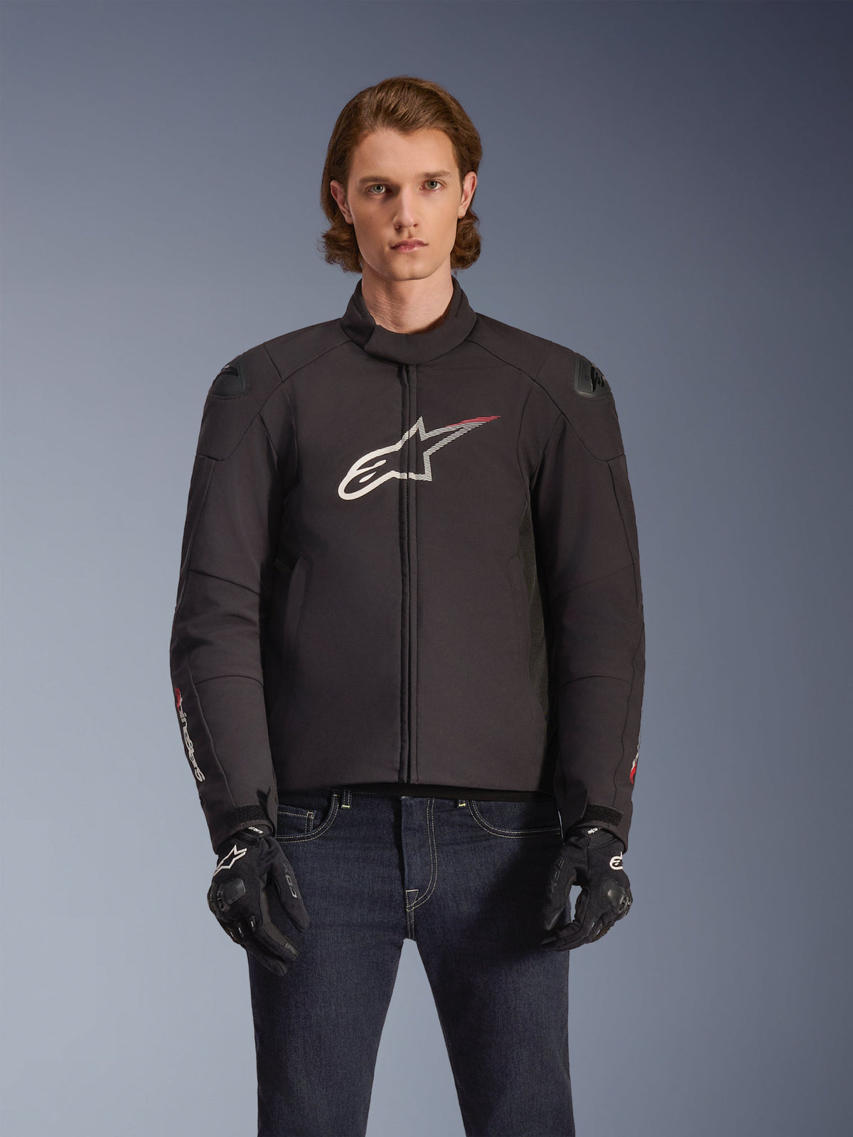 Alpinestars SMX Waterproof Jacket, Black textile motorcycle jacket with white and red star logo on the chest, worn by a model with black gloves and blue jeans, featuring external shoulder sliders and a sporty fit.