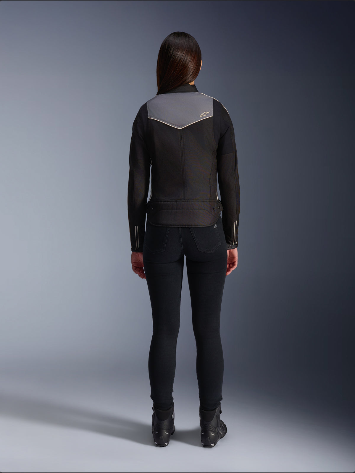 Rear view of a female rider wearing the Alpinestars Stella T-Dyno WR Jacket in Black, Dark Gray, and Ecru, paired with black slim-fit pants and black motorcycle boots, highlighting the gray upper back panel with Alpinestars logo and ecru trim details.