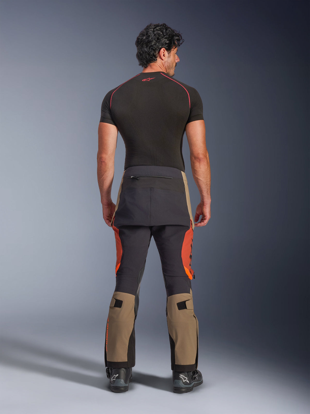 Alpinestars Halo Pro Drystar® XF Laminated Pants, Walnut Rust Black adventure motorcycle pants, back view on a model wearing black boots and a short-sleeved base layer, featuring walnut-colored lower legs, rust orange accents, and black reinforced panels.