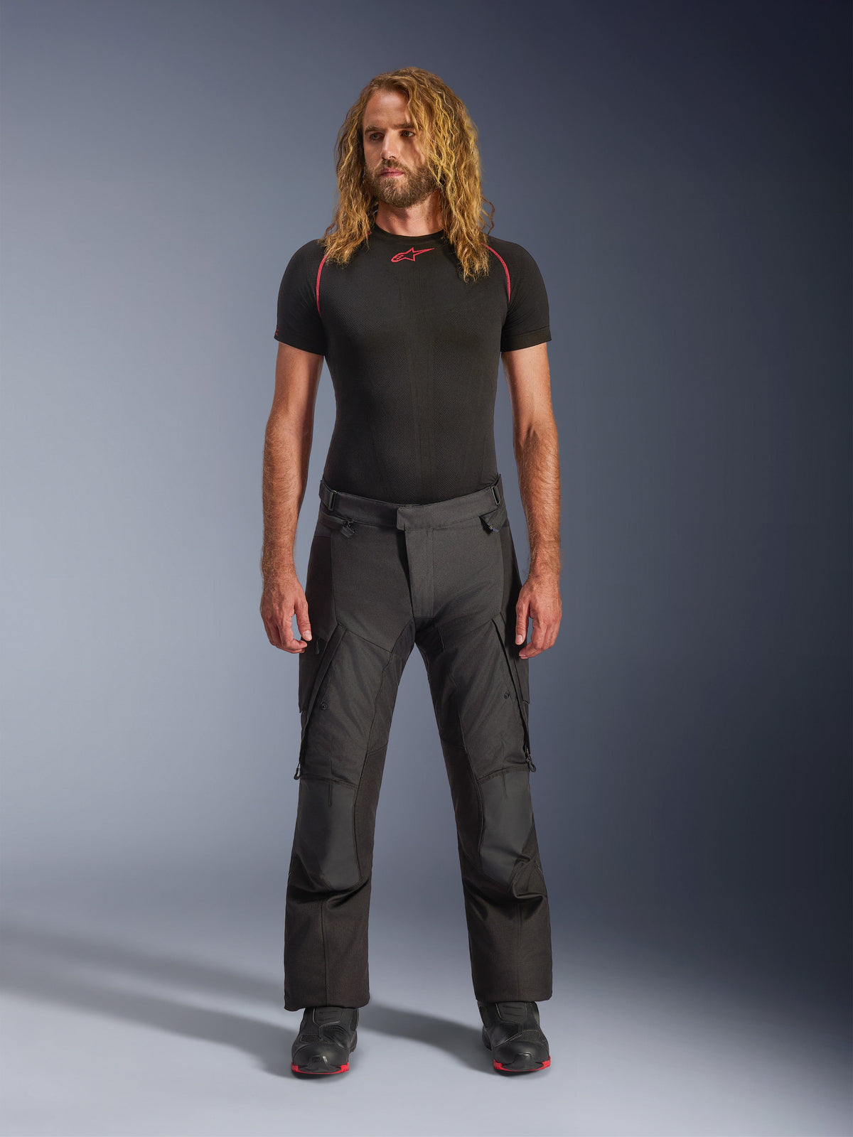 Male model wearing Alpinestars Andes V4 Drystar® Pants - Long in Black, adventure-touring motorcycle pants featuring reinforced knee panels and cargo pockets, styled with a black short-sleeve base layer and black motorcycle boots.