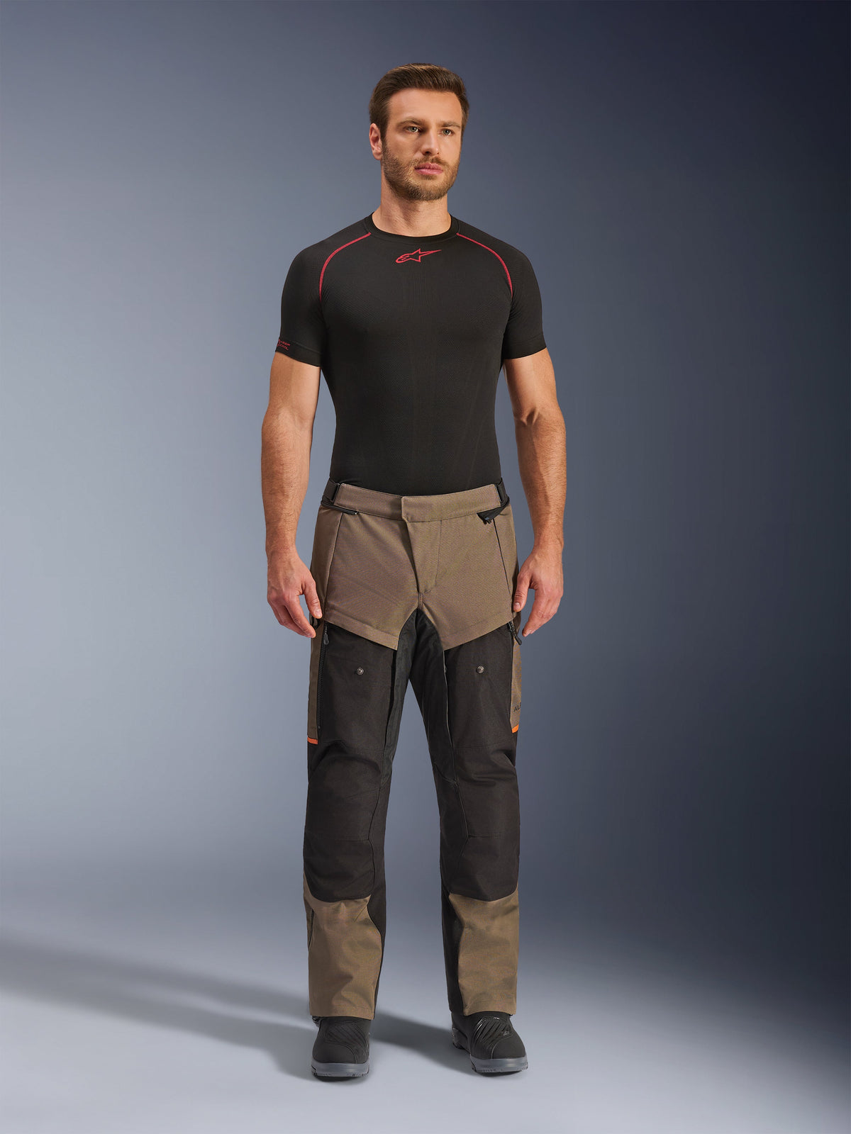 Alpinestars Cusco Drystar® Pants, Walnut Black, adventure touring motorcycle pants, worn by a male model with a black base layer and black boots, featuring walnut and black panels, orange highlights, and direct ventilation zippers on the thighs.