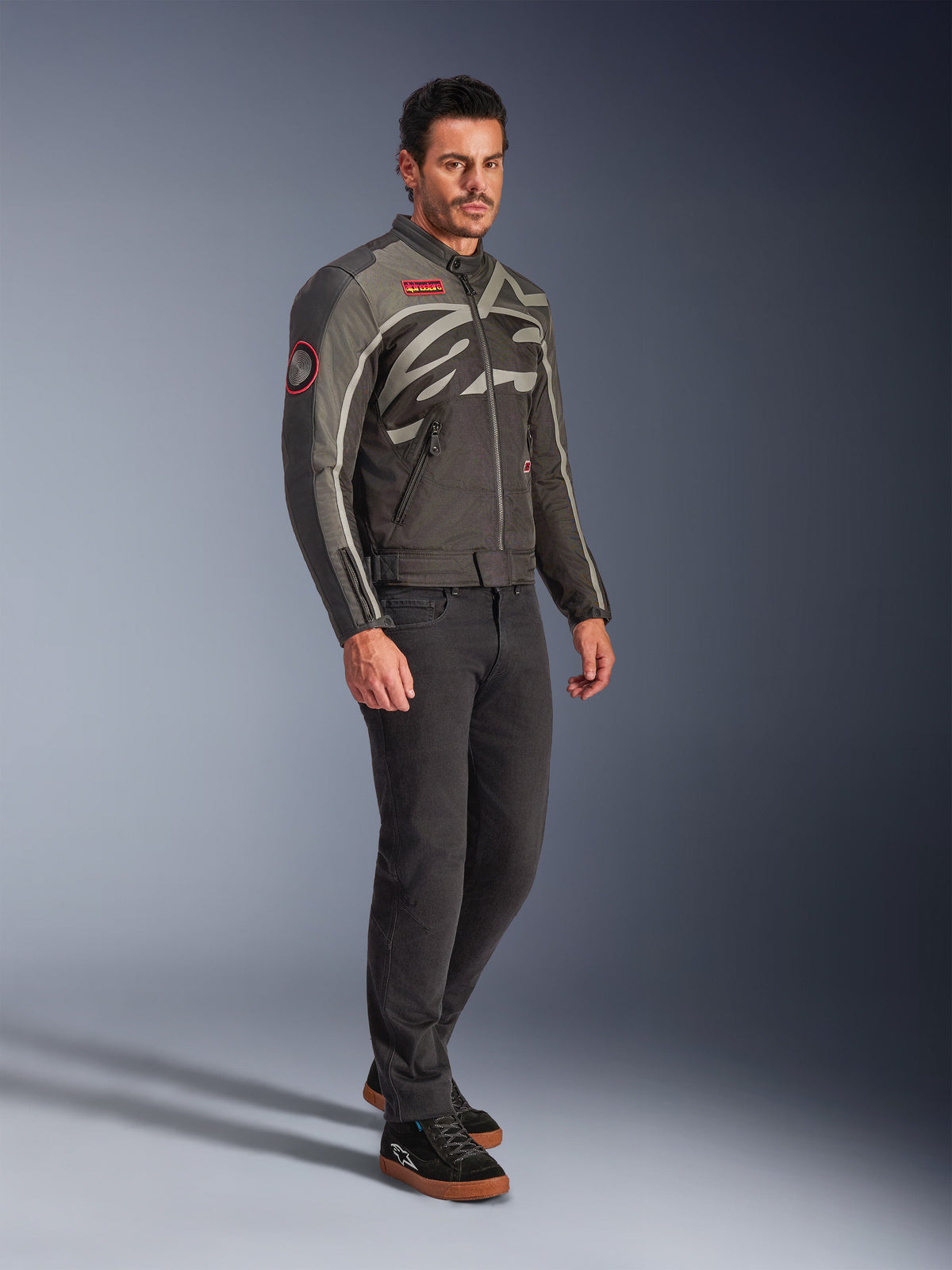 Male model wearing the Alpinestars Axton Jacket in Black Anthracite Red Tangerine, vintage-inspired motorcycle jacket with a wrap-around logo and red accents, paired with black pants and black riding sneakers, urban contemporary motorcycle gear