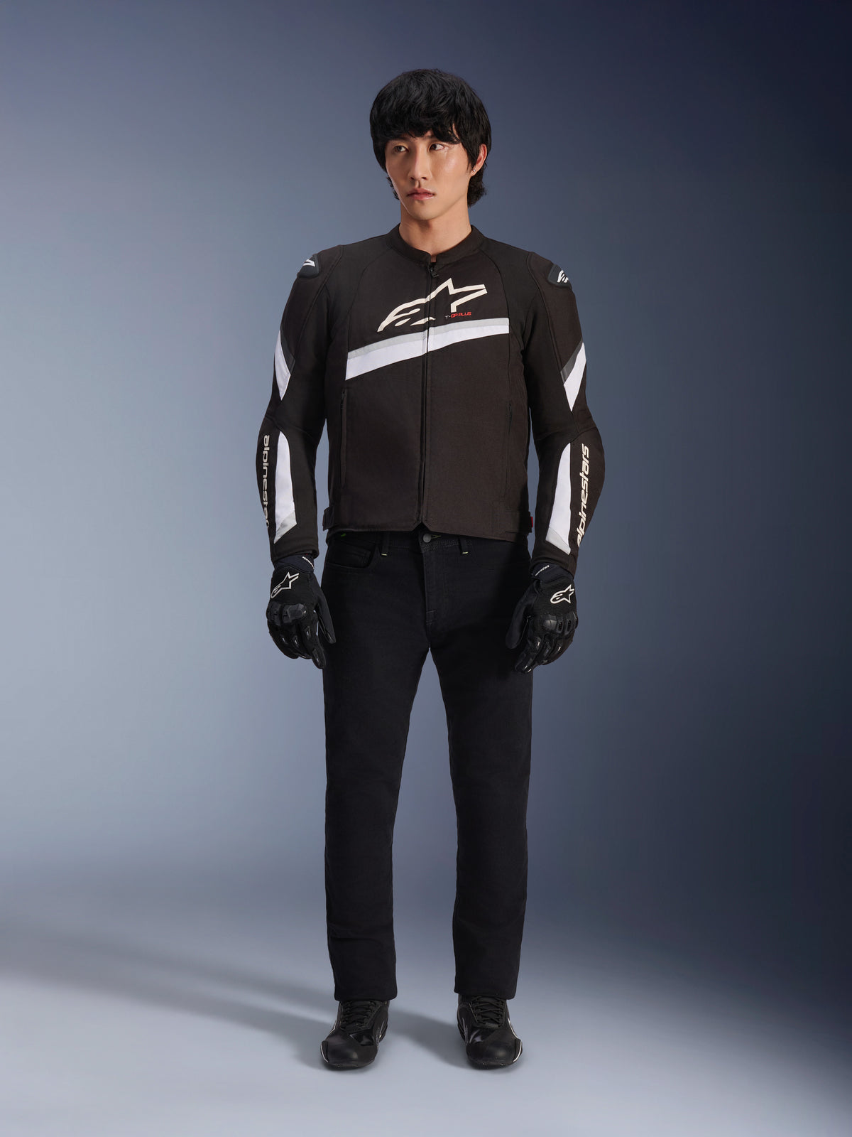 Alpinestars T-GP Plus R V4 Jacket, Black and White, Sport Textile Motorcycle Jacket, worn by a model with black pants and gloves, featuring bold white Alpinestars logo on the chest and sleeves, sleek racing-inspired design