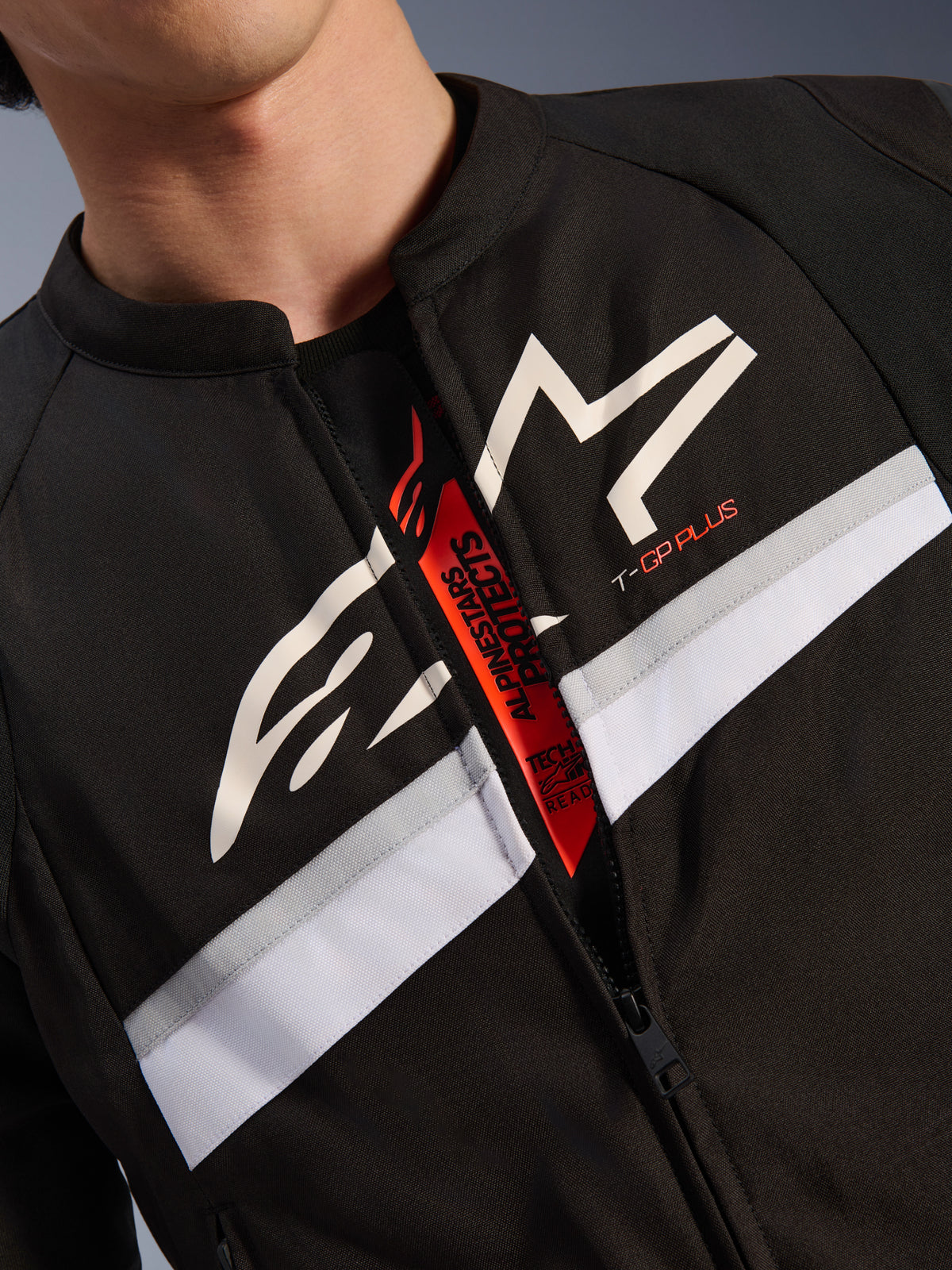 Alpinestars T-GP Plus R V4 Jacket, close-up of chest and neck, black textile with white star logo and T-GP PLUS branding, white and grey horizontal stripe, open zipper revealing red interior tab with Tech-Air Ready text
