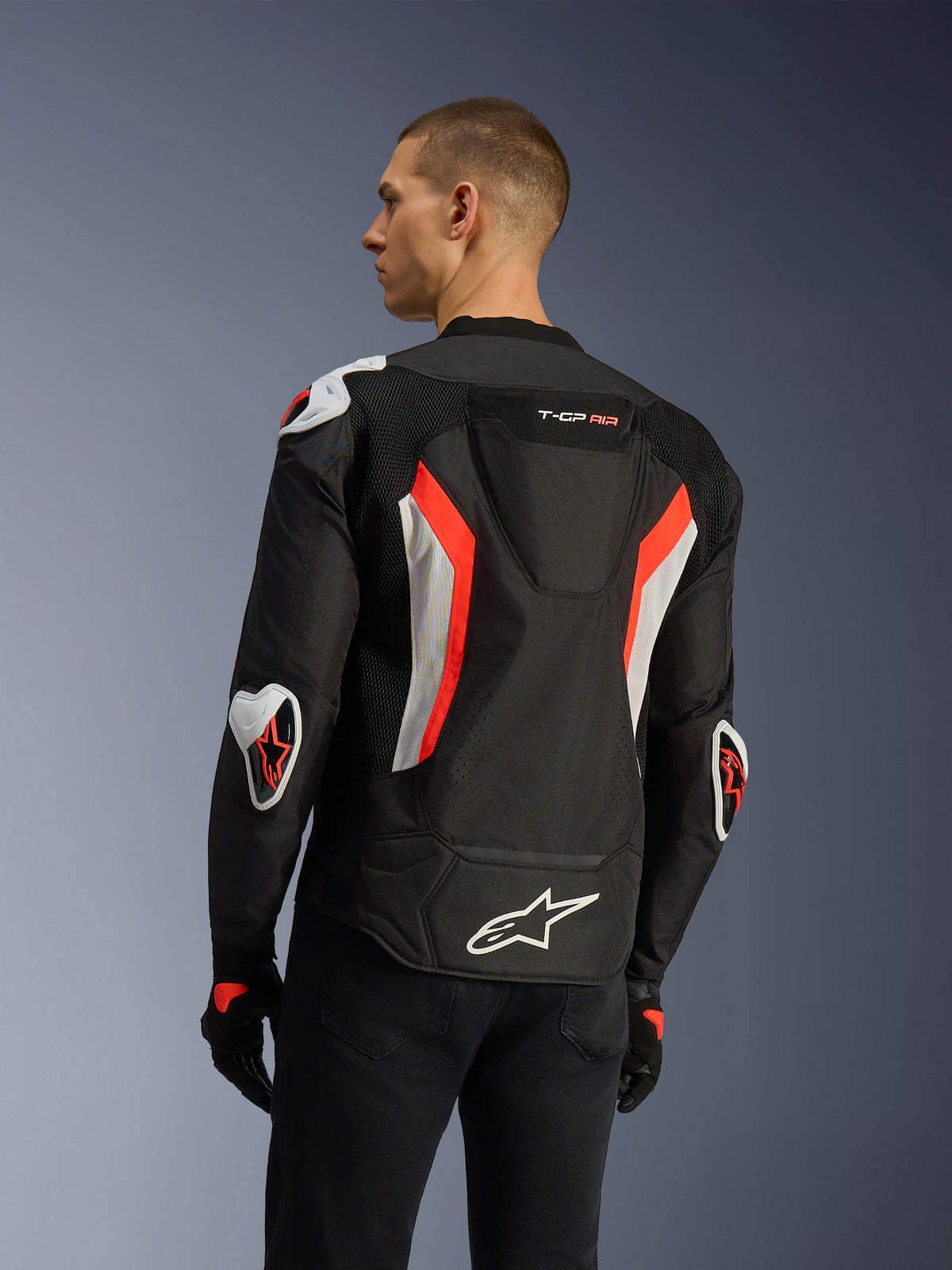 Alpinestars T-GP Air Jacket in Black Red Fluo, rear three-quarter view on a model, highlighting the aerodynamic hump, Racing GP DFS external armor on shoulders and elbows, laser-perforated mesh panels, and white and red fluo accents, sport-cut textile motorcycle jacket.
