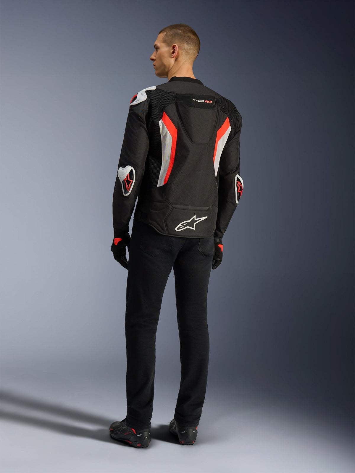 Alpinestars T-GP Air Jacket, Black Red Fluo motorcycle jacket, rear view on a model, featuring mesh ventilation panels, aerodynamic hump, and white external protectors on elbows and shoulders, worn with black pants and riding shoes
