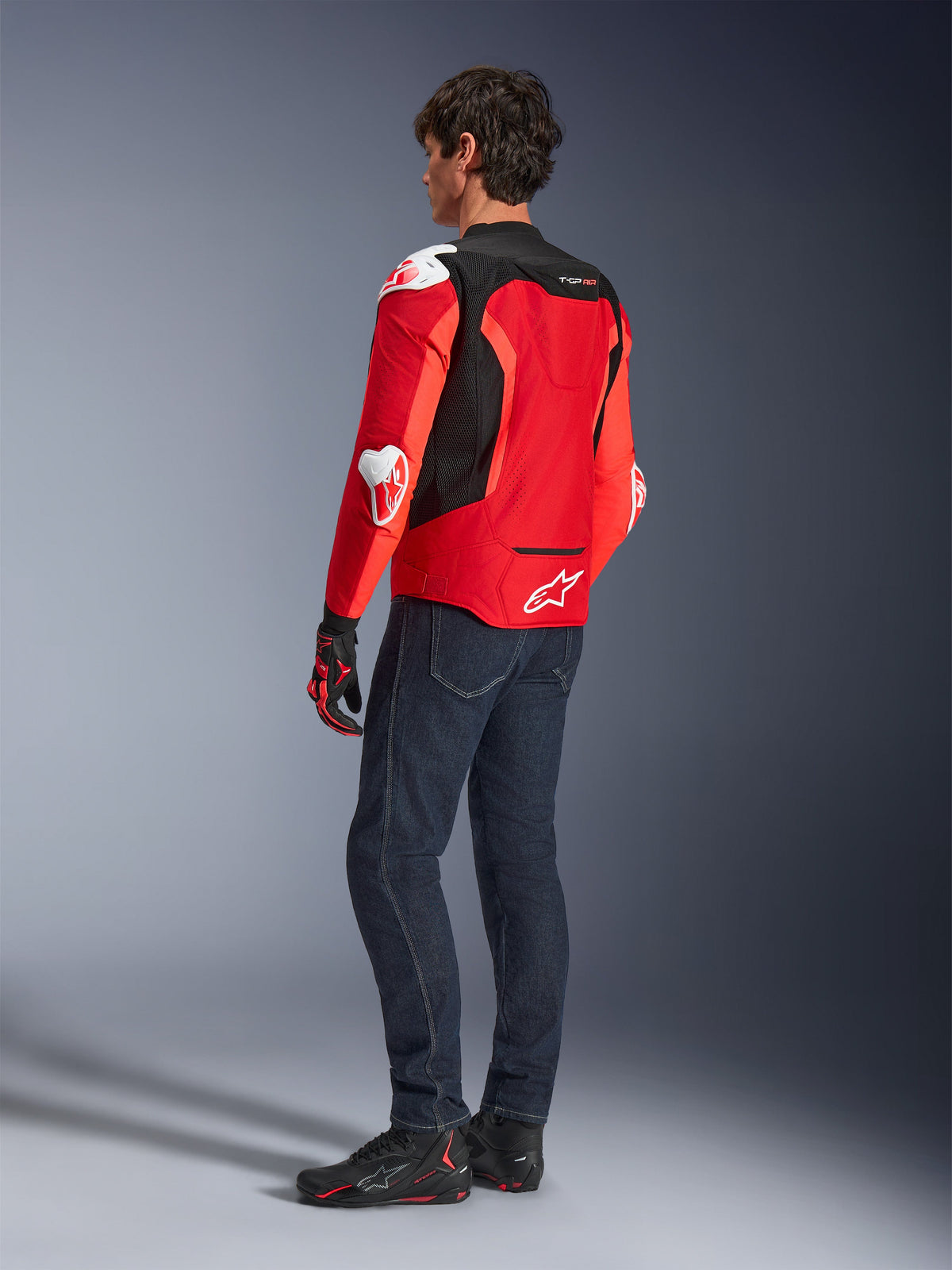 Alpinestars T-GP Air Jacket, Rear view of a man wearing a bright red and fluo red textile jacket with black mesh panels and white armor sliders, paired with dark blue jeans and black riding shoes, sport motorcycle riding gear.
