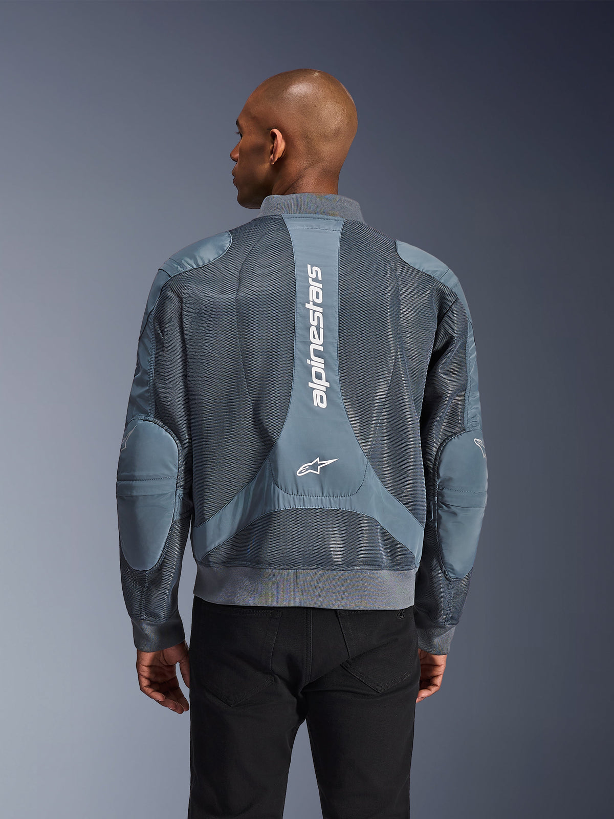 Alpinestars Flight Air Jacket in Storm Gray, rear view on a model, urban motorcycle jacket featuring extended 3D mesh panels, vertical Alpinestars branding on the back, and a bomber-style ribbed hem and collar.