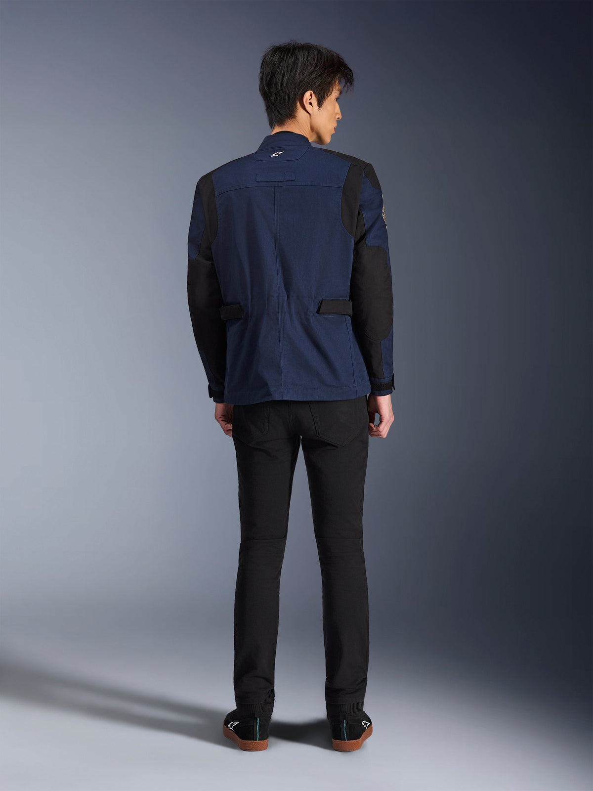 Model wearing the Alpinestars Dice Jacket in Dark Blue and Black, back view, urban motorcycle field jacket featuring box-pleated shoulders and adjustable waist tabs, paired with black pants and black sneakers.
