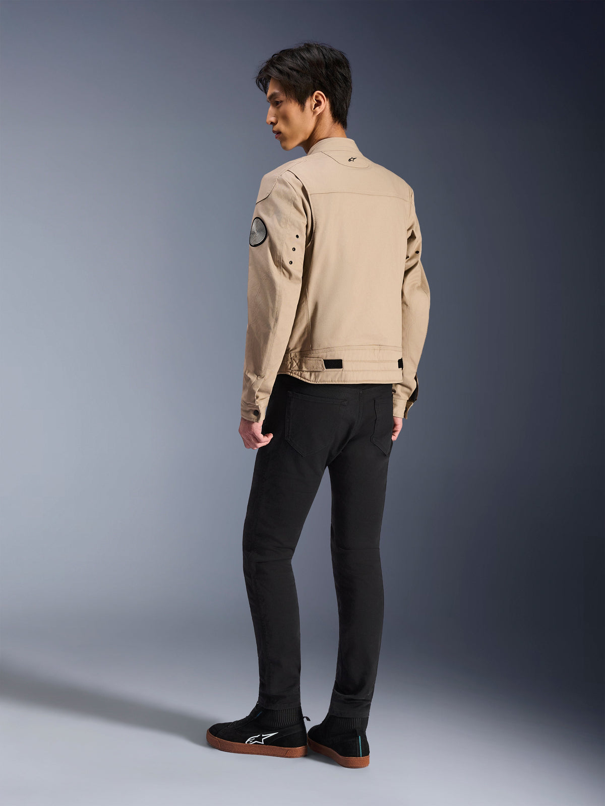 Male model from the back wearing the Alpinestars Bruiser Jacket in tan, urban contemporary motorcycle jacket with workwear styling, featuring a circular sleeve patch, waist adjustment straps, and articulated shoulders, paired with black pants and black sneakers.