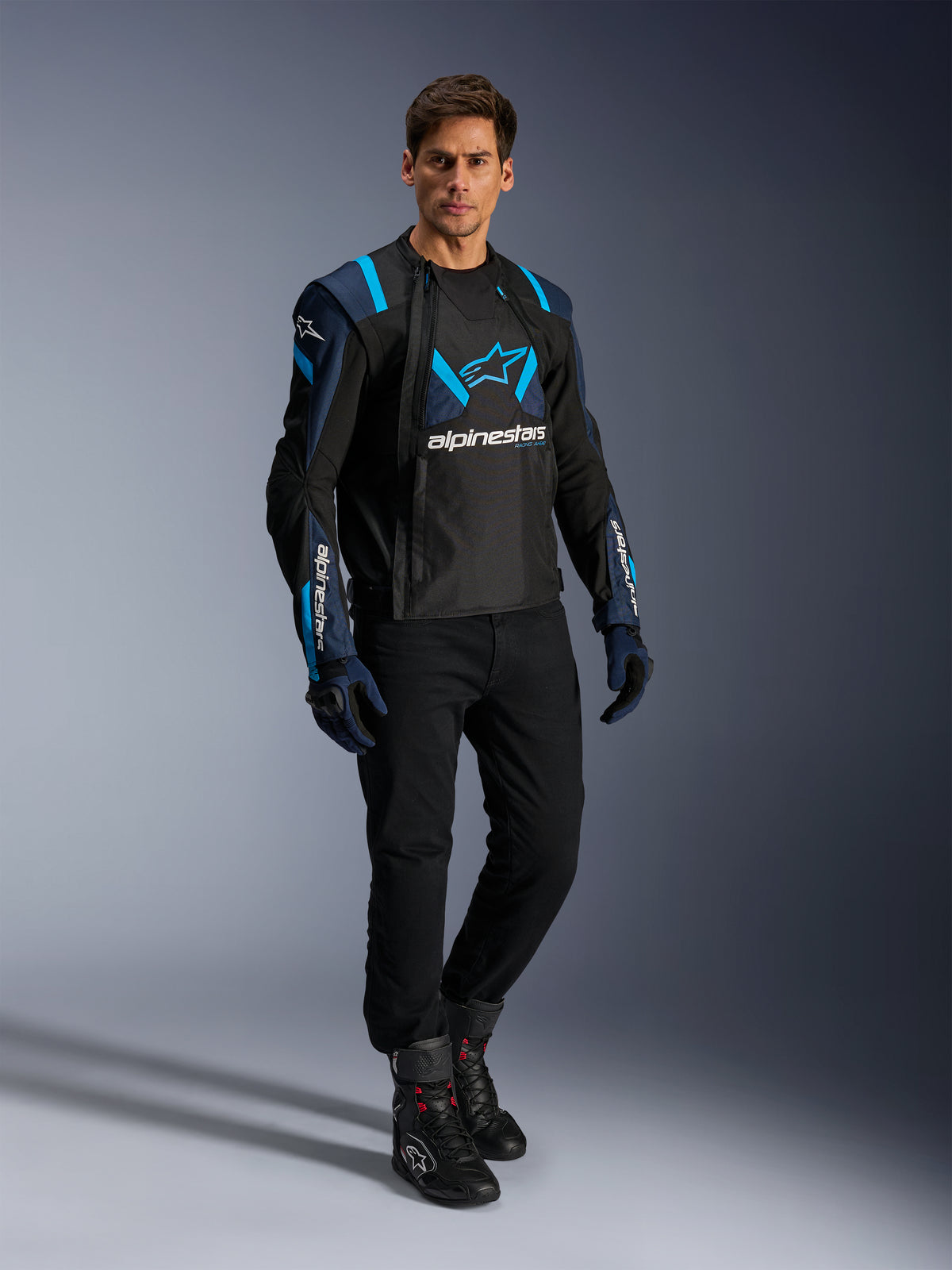 Male model wearing Alpinestars T-Stunt Air Jacket, Black Dark Blue and Blithe Blue, featuring prominent mesh panels for airflow, paired with black pants and riding gloves, sport-cut motorcycle jacket for summer road riding.