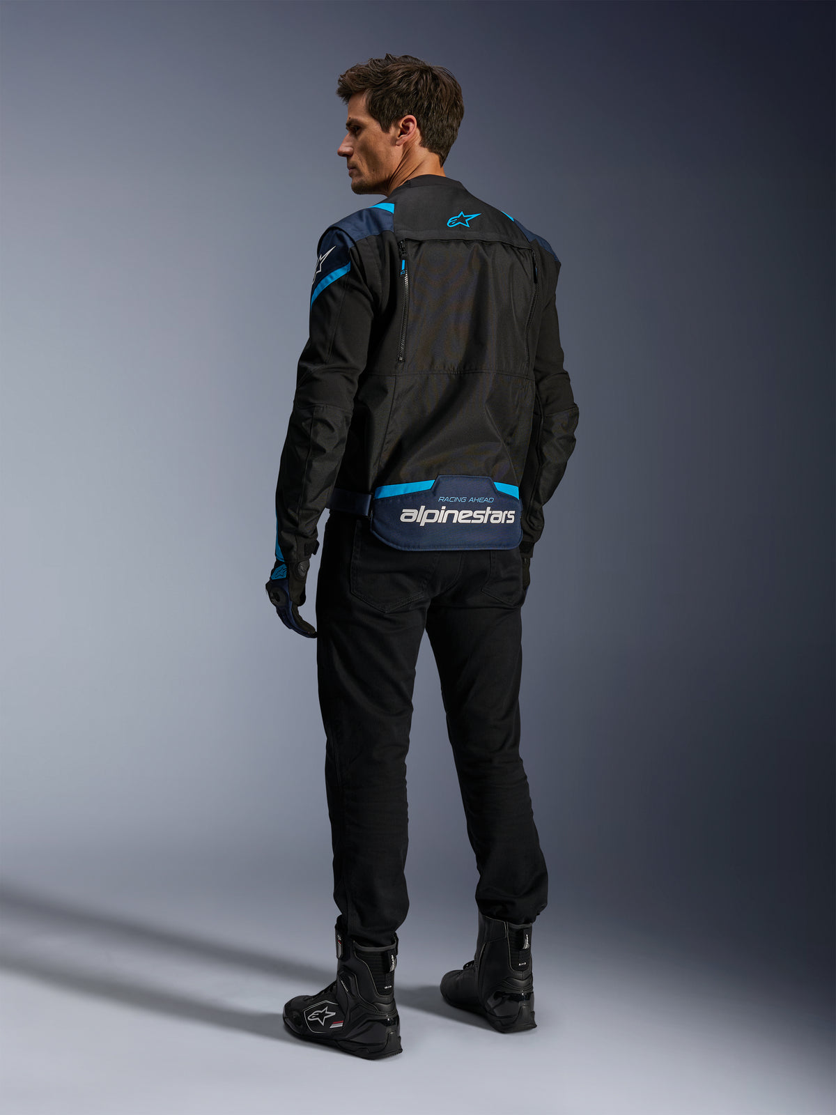 Alpinestars T-Stunt Air Jacket, Motorcycle Jacket, Black with Dark Blue and Blithe Blue accents, rear view showing modular mesh panels and branding on the lower back, worn by a model with black riding pants and boots.