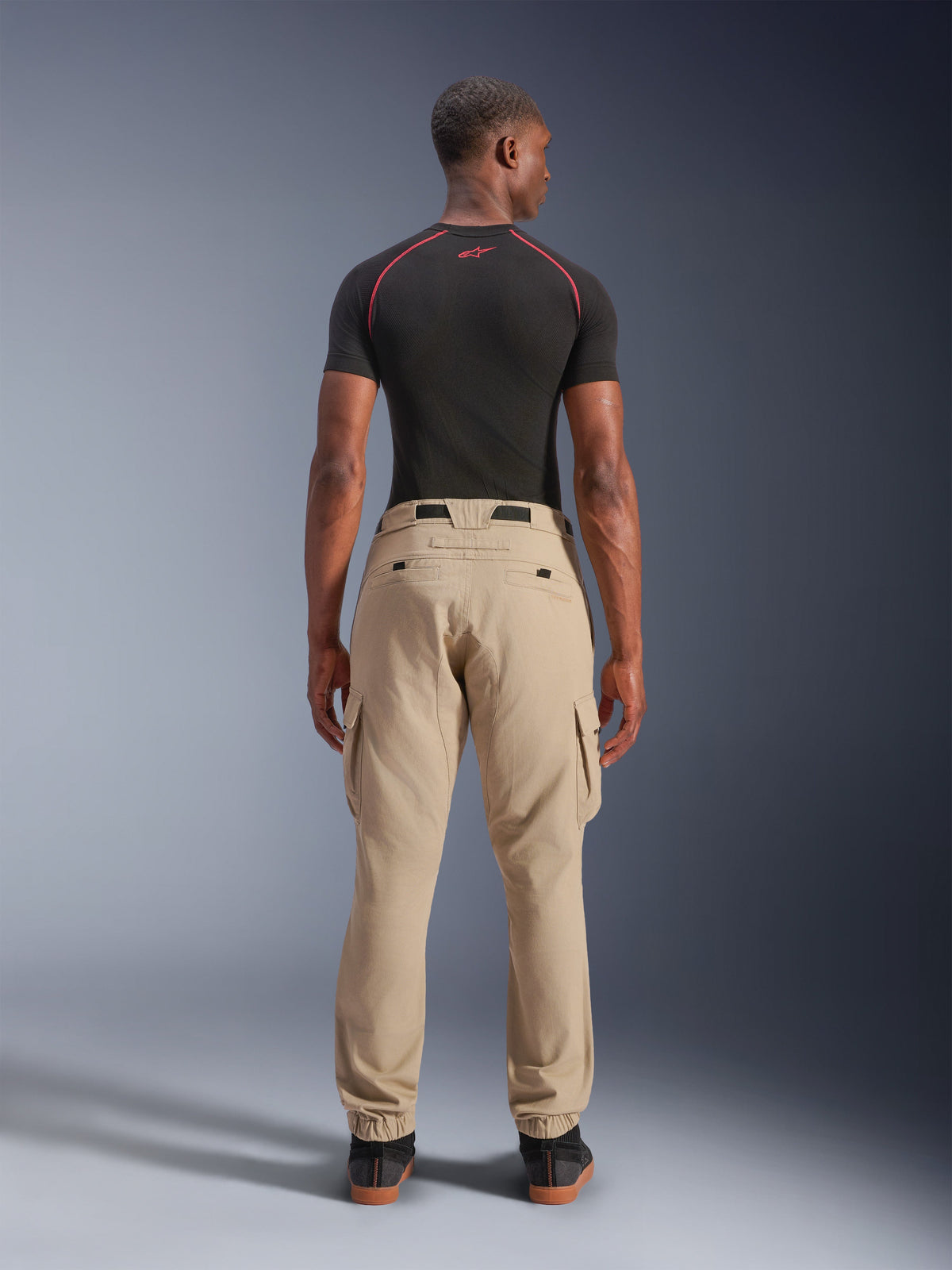 Alpinestars Flex-AST Cargo Pants - Short, Light Beige, back view on a model, regular tapered fit with elasticated hems and cargo pockets, worn with a black technical base layer with red accents, modern urban motorcycle apparel