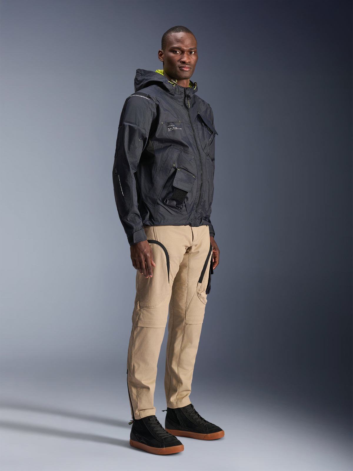 Alpinestars Acteon Pants, urban motorcycle pants, Sand color, worn by model with a dark technical jacket and black sneakers, featuring a tapered fit with black zipper accents and cargo pockets, contemporary motorsport-inspired design