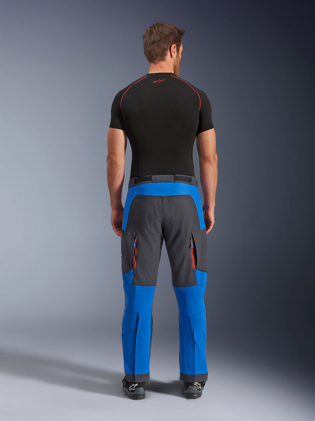 Alpinestars Maxdura Dual Pants in Royal Blue and Deep Gray, adventure motorcycle pants shown from the back, featuring zippered cargo pockets with orange accents and an over-the-boot design, worn with a black short-sleeve base layer