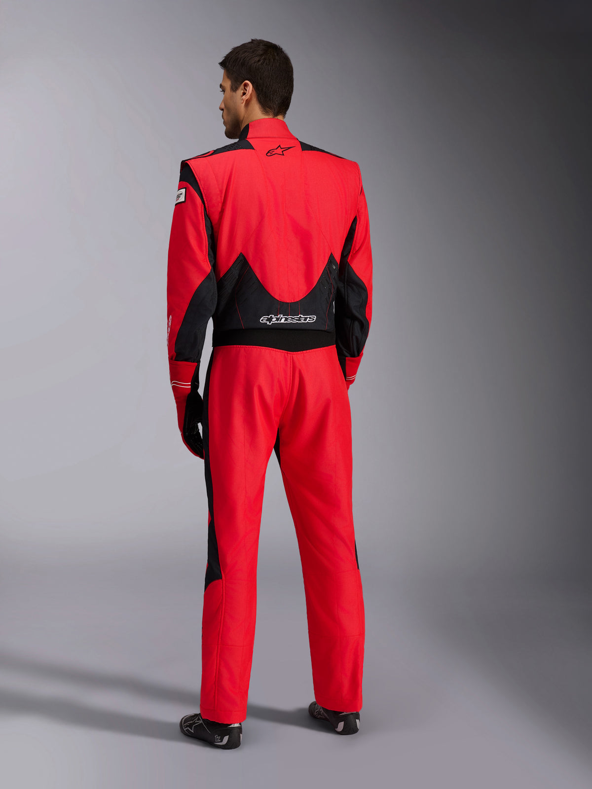 Full back view of Alpinestars GP Pro Comp V2 FIA Racing Suit, Red and Black, featuring breathable black mesh panels on the shoulders and lower back, white Alpinestars logo, worn with matching racing gloves and boots, professional motorsport driver gear