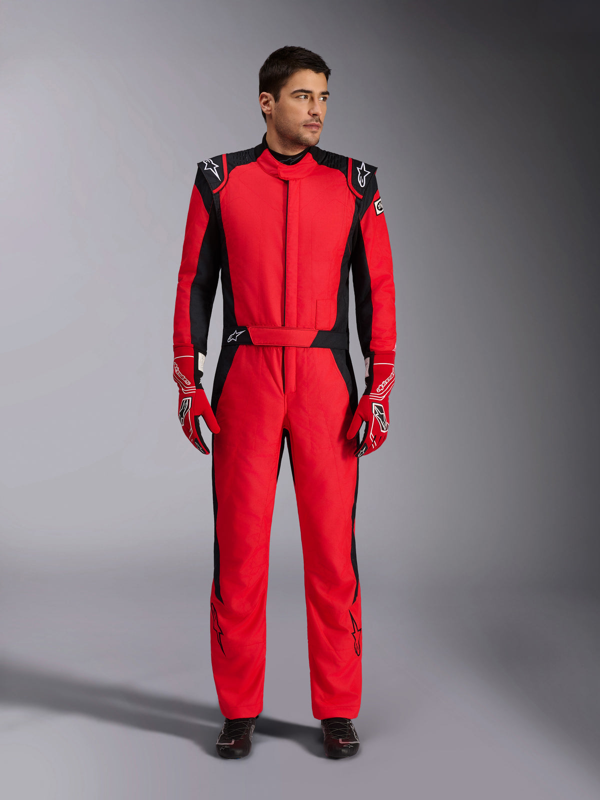 Male model wearing Alpinestars GP Pro Comp V2 FIA Auto Racing Suit, Red and Black, paired with red racing gloves and black shoes, professional one-piece motorsport suit featuring shoulder epaulettes and Alpinestars branding, high-performance driver gear for auto racing