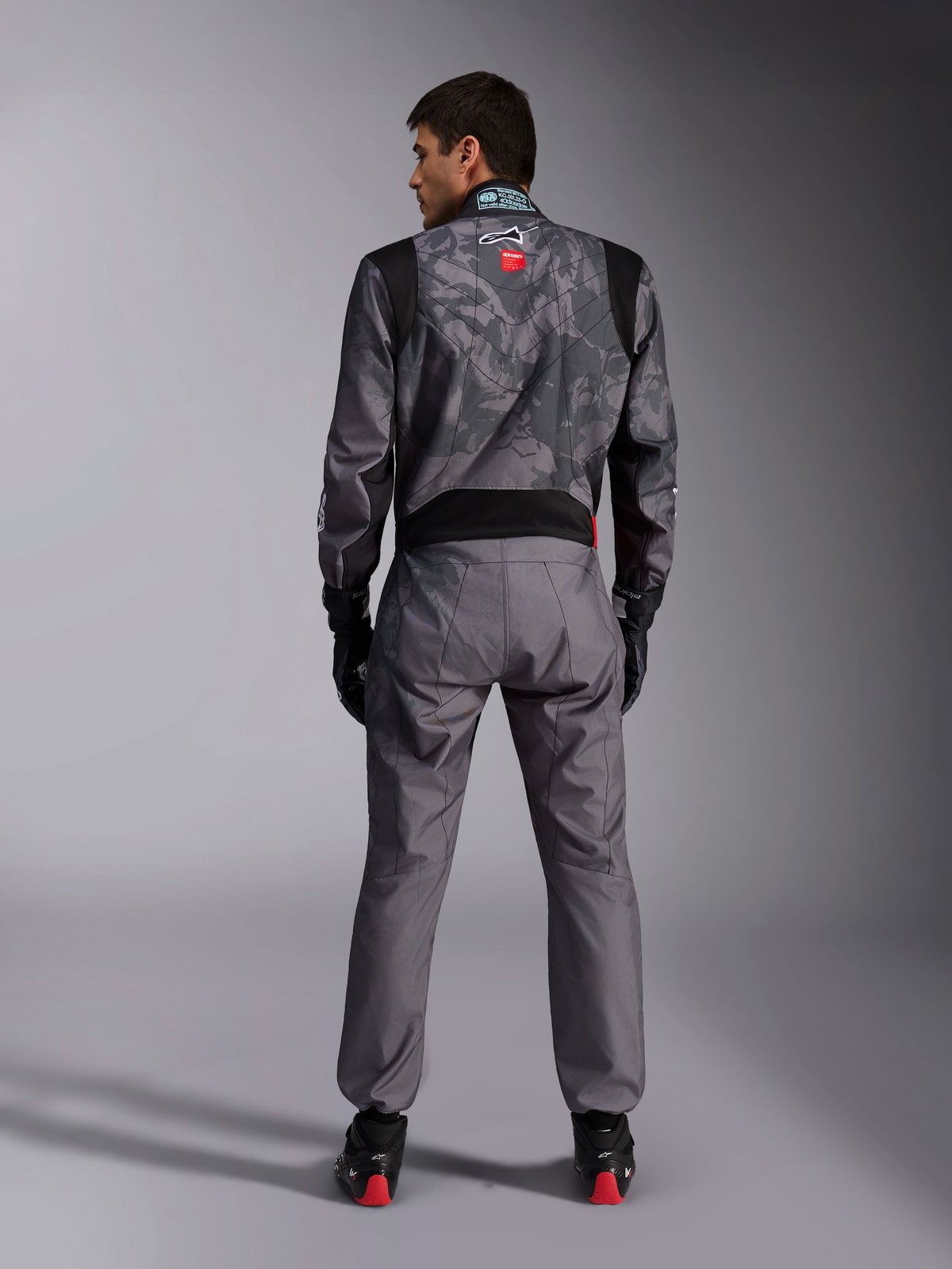 Alpinestars KMX-9 V3 Graphic 5 Suit, Karting Suit, Dark Gray and Black camouflage pattern on the upper body, back view showing stretch panels and Alpinestars branding, worn with black gloves and black racing shoes with red details, professional motorsport racing gear