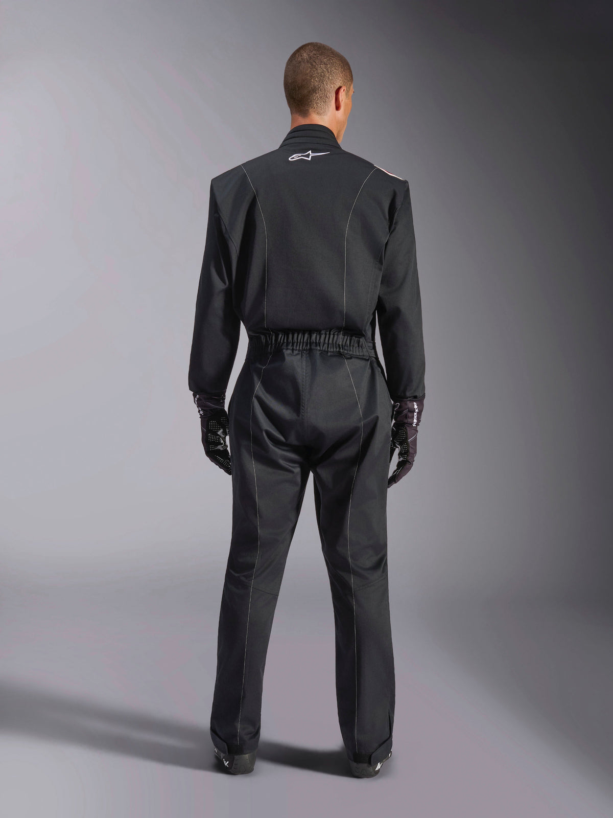 Alpinestars Kart Indoor V2 Suit, Black Karting Suit, rear full-body view on model, featuring white piping details and a small white Astars logo on the back collar, elastic waist belt, worn with black racing gloves, one-piece motorsport apparel.