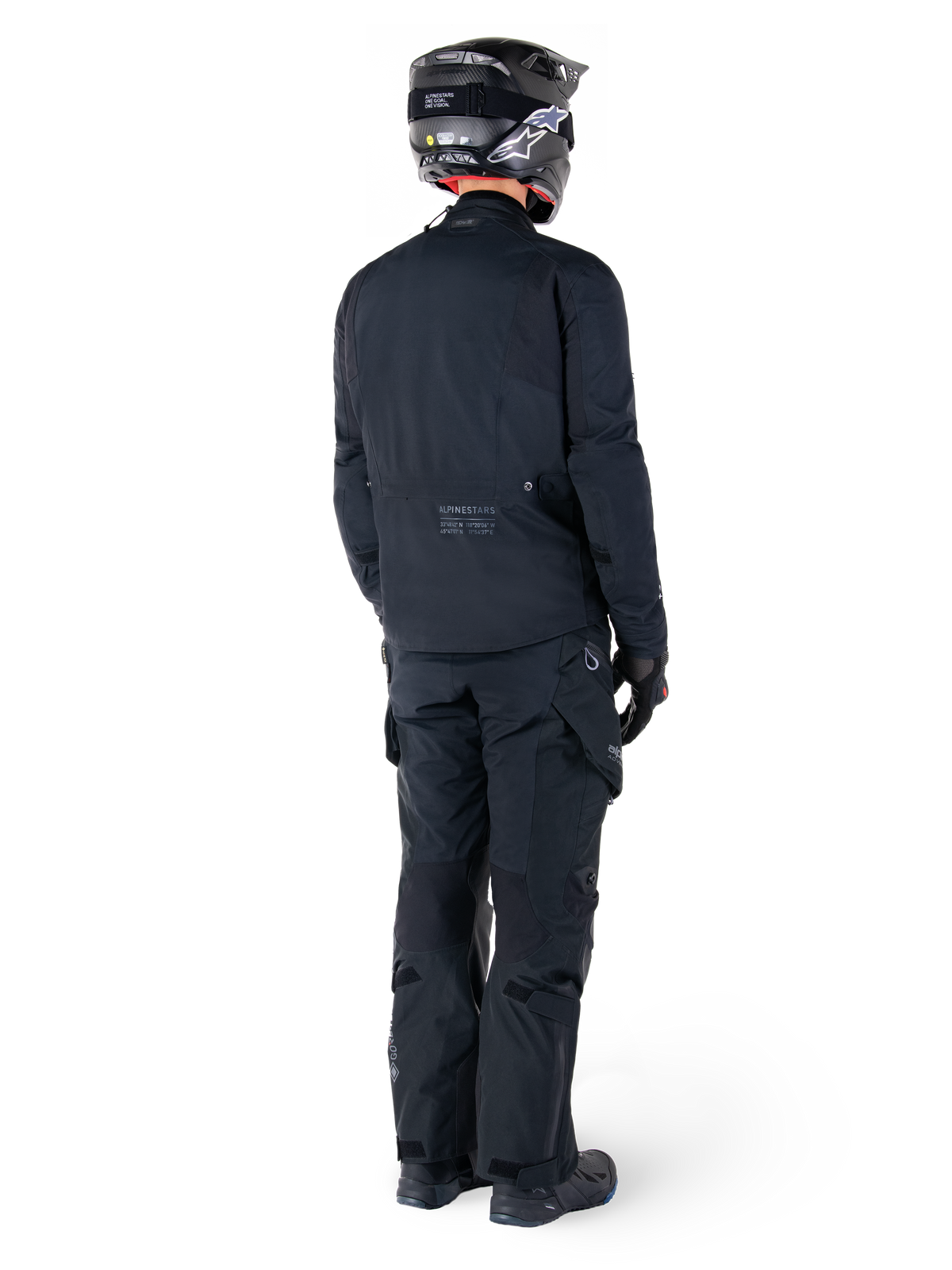 Rear view of a rider wearing the Alpinestars St-7 Prime GORE-TEX Jacket in black, paired with matching black touring pants, a black motorcycle helmet with white logo, and black riding boots, full protective adventure gear for touring and commuting.