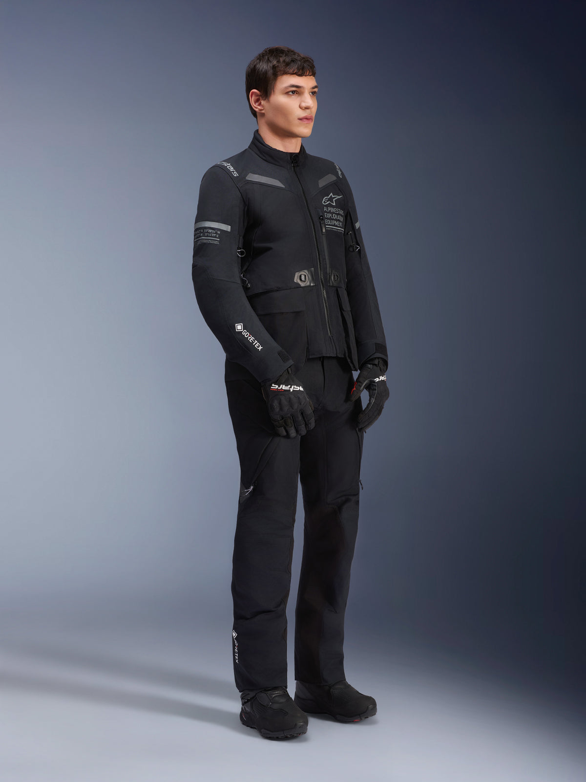 Male model wearing Alpinestars ST-7 2L GORE-TEX Jacket in Black Dark Gray, adventure touring motorcycle jacket, matching black pants and gloves, three-quarter full-body view, featuring GORE-TEX logo and Alpinestars Exploration Equipment branding