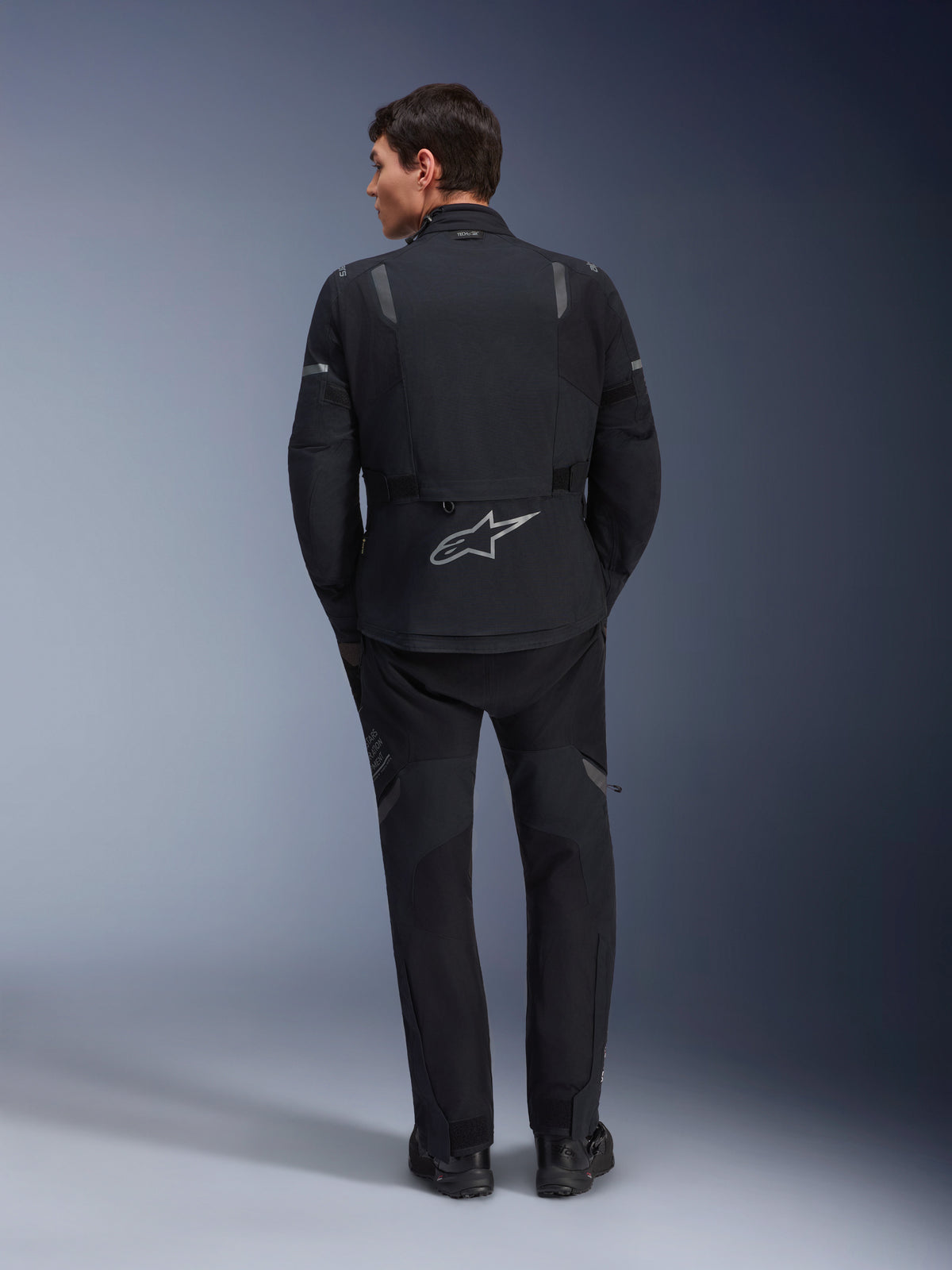 Alpinestars ST-7 2L GORE-TEX Jacket, Adventure Motorcycle Jacket, Black and Dark Gray, rear view of a man wearing the jacket and matching black pants, featuring a large reflective Astar logo on the lower back, waterproof textile gear for touring and road riding