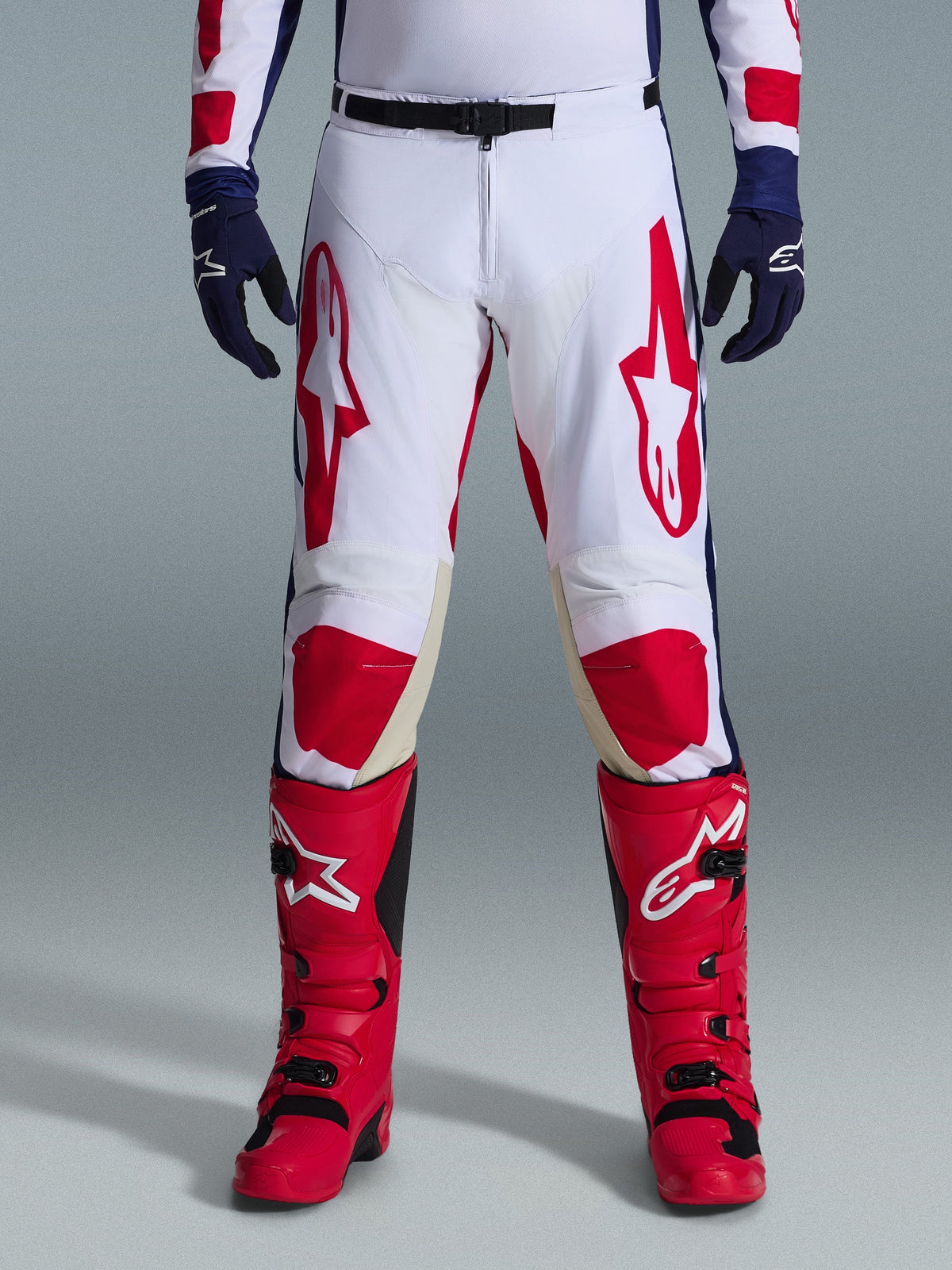 Alpinestars 2026 Racer Riway Pants, Motocross Pants, White Blue and Red, worn with red boots and navy gloves, featuring large red logos on the thighs and red knee panels, sporty pre-curved design with adjustable waist buckle
