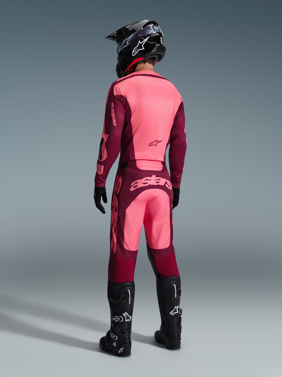 Motocross rider wearing the Alpinestars 2026 Racer Riway Gear Set from a rear view, maroon and light pink colorway, jersey and pants with prominent branding, complete with black helmet, gloves, and boots, coordinated motocross racing outfit