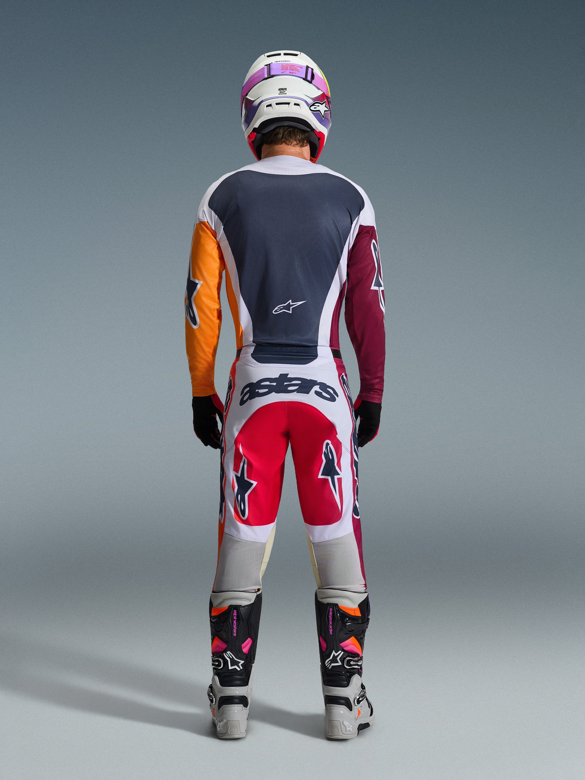 Motocross rider viewed from behind wearing the 2026 Racer Portl Pants in Red, Light Gray, and Dark Gray, featuring "astars" branding across the seat, coordinated with a multi-colored jersey, white helmet, and technical racing boots.