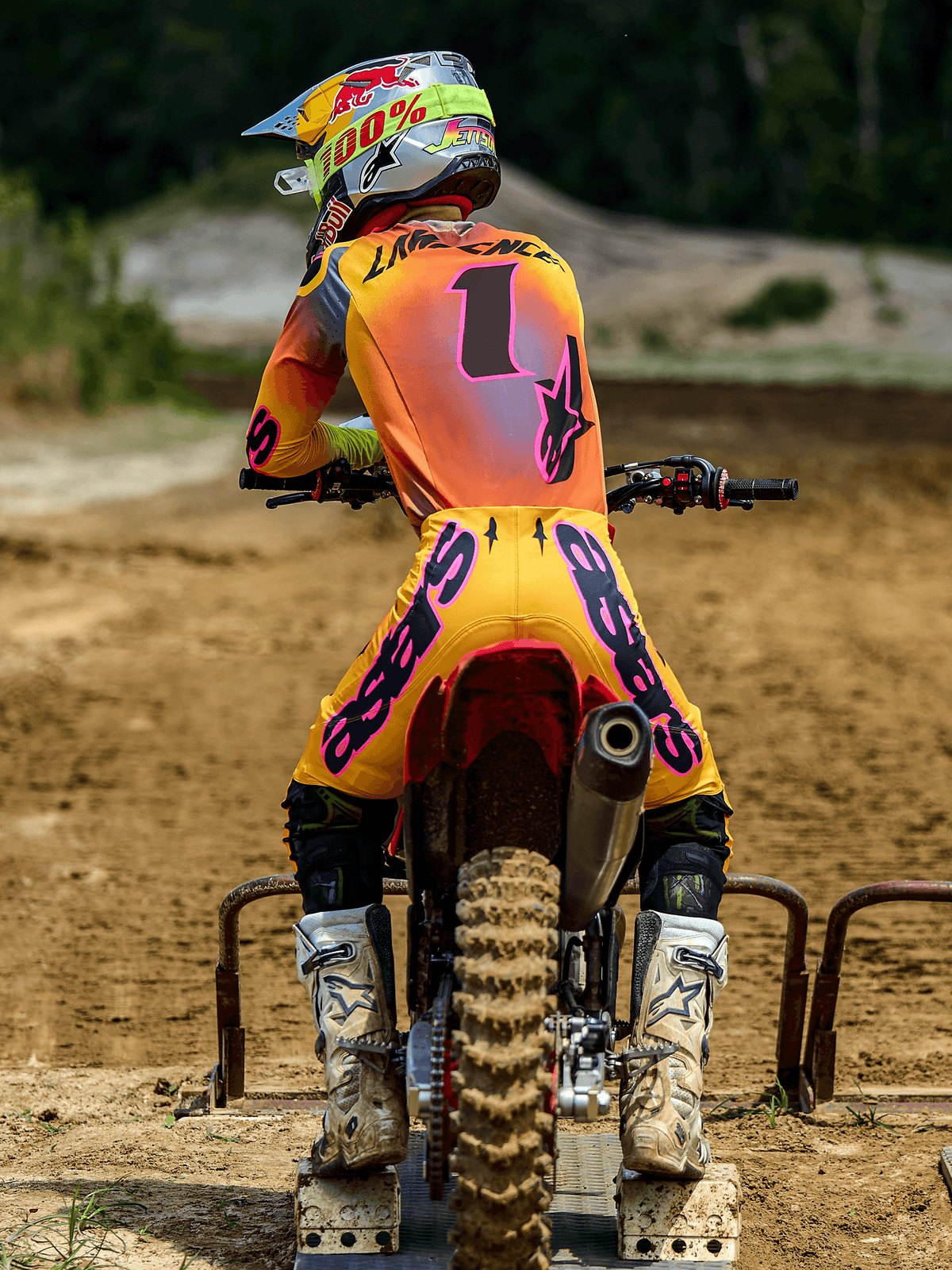 Motocross rider from behind wearing Alpinestars 2025 Supertech Lipan Gear Set, Yellow and Pink, jersey with the number 1 and 'LAWRENCE' on the back, yellow pants with bold pink 'ASTARS' branding, white boots and matching helmet, seated on a dirt bike at the starting gate