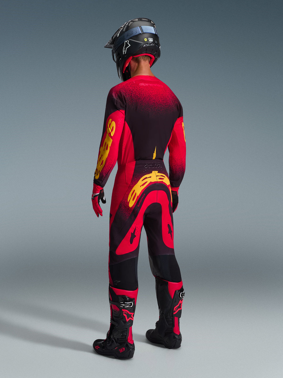 Motocross rider seen from behind wearing Alpinestars 2026 Supertech Scenz Pants in Red Black Yellow, matching jersey with bold yellow branding, black and red motocross boots, black helmet, complete professional racing gear set for off-road performance.