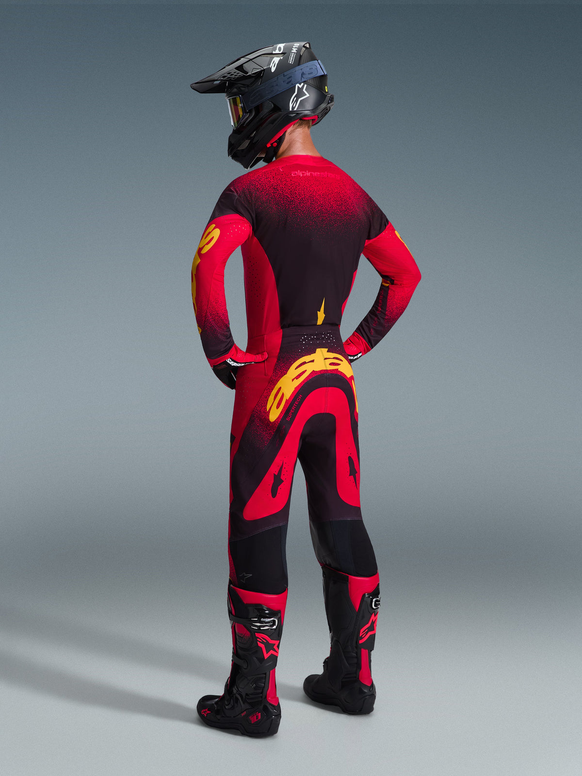 Motocross rider from behind wearing Alpinestars 2026 Supertech Scenz Pants in Red Black Yellow, featuring a large yellow logo on the seat, paired with a matching red and black gradient jersey, black helmet, and red motocross boots, professional racing gear set for off-road performance.
