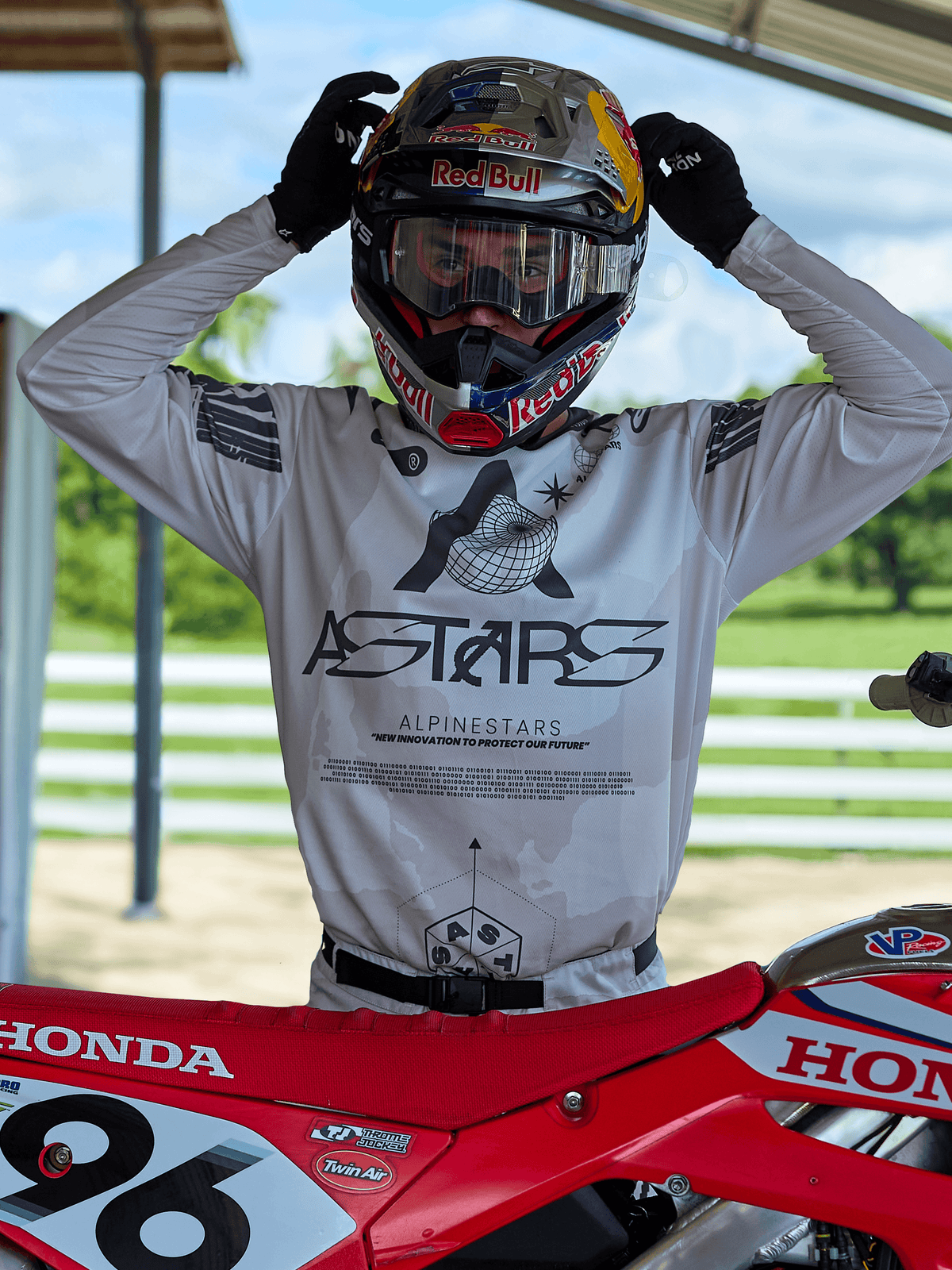 Motocross rider wearing Alpinestars 2025 Racer Hollow gear, light camo jersey with bold ASTARS branding, adjusting a Red Bull helmet with clear goggles, standing next to a red Honda dirt bike in an outdoor off-road setting
