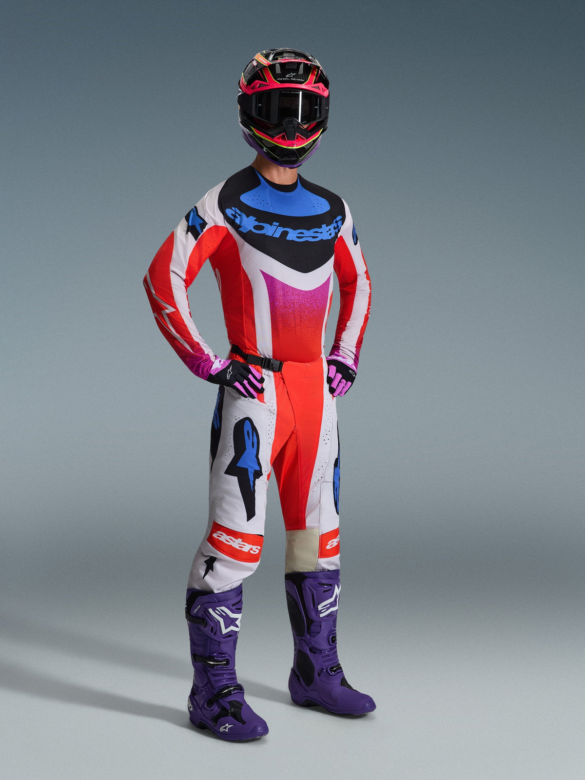Motocross rider wearing Alpinestars 2026 Techstar Knif Pants, Light Gray and Orange Fluo with blue accents, paired with a matching multi-colored jersey, purple off-road boots, and a colorful racing helmet, complete professional racing gear set.
