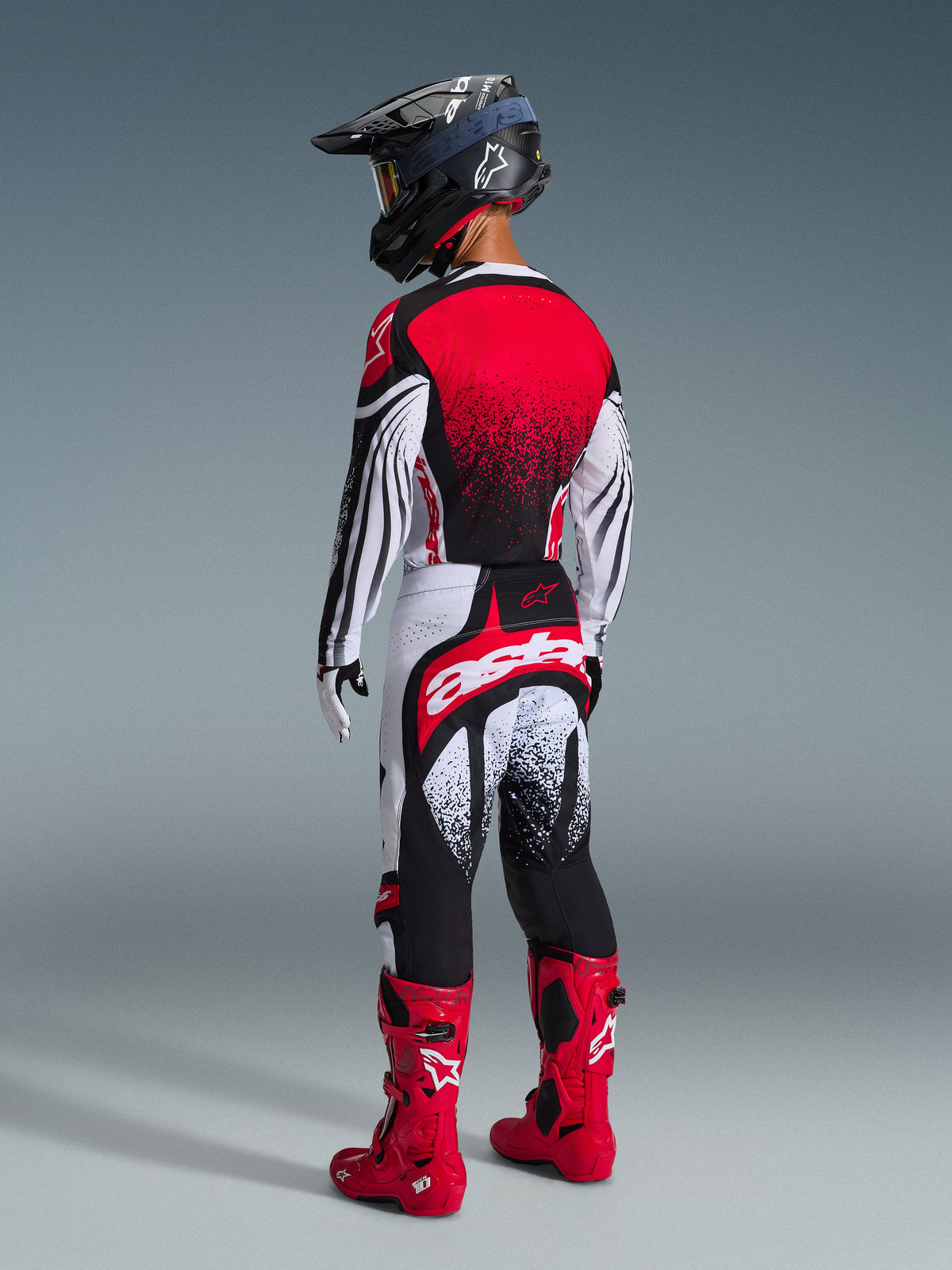Motocross rider seen from behind wearing the Alpinestars 2026 Techstar Nomur Pants in Black White and Red, featuring a large white 'astars' logo across the seat, paired with a matching jersey, bright red motocross boots, and a black helmet, professional gear set for off-road racing