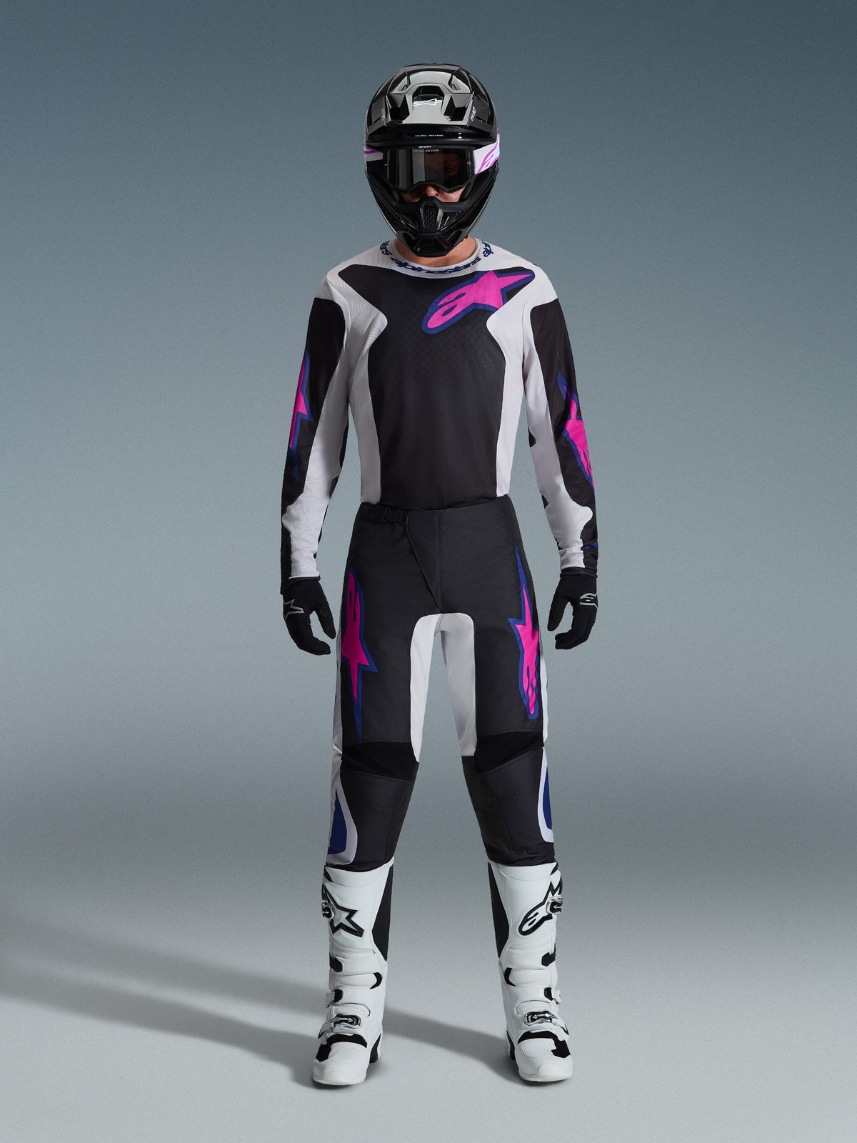 Motocross rider wearing the Alpinestars 2026 Fluid Grid Gear Set, black and light gray jersey with bright pink logos, black and white pants with matching pink and blue graphics, black helmet with pink goggles, white and black boots, full off-road racing gear ensemble