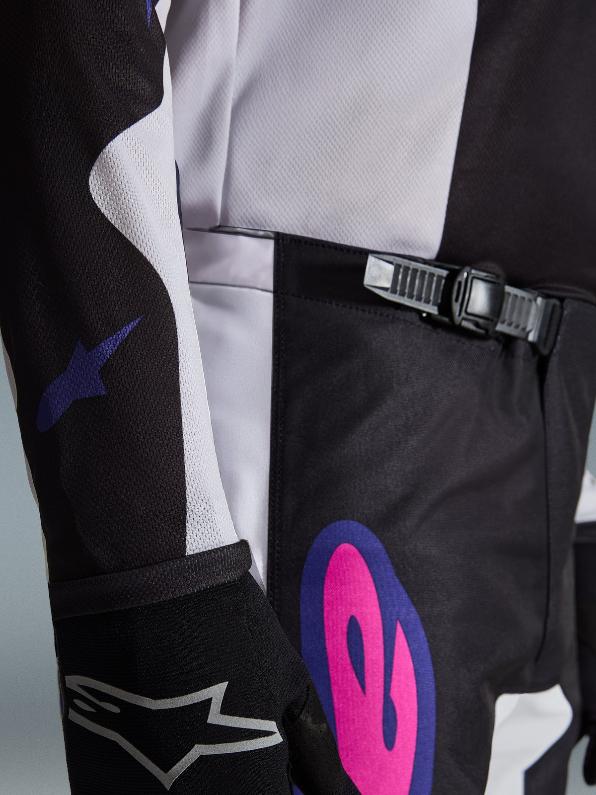 Alpinestars 2026 Fluid Grid Pants, Motocross Pants, Black Light Gray and Purple, close-up featuring the ratchet closure system at the waist and a bold pink and purple graphic on the hip, worn with a matching jersey and black glove.