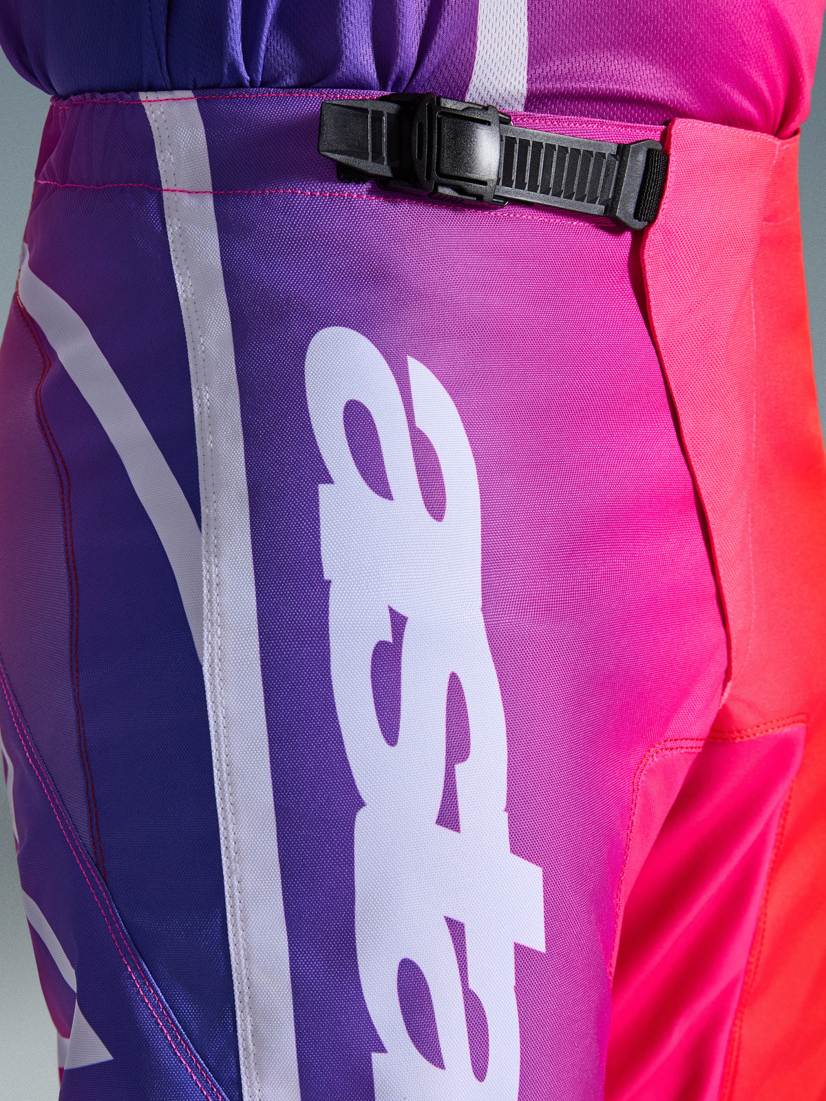 Alpinestars 2026 Fluid Apex Pants, Motocross Pants, Multicolor Purple Pink and Red, close-up of the waist and hip area featuring a black ratchet closure system and large white vertical Alpinestars logo, vibrant gradient design with durable polyfabric texture