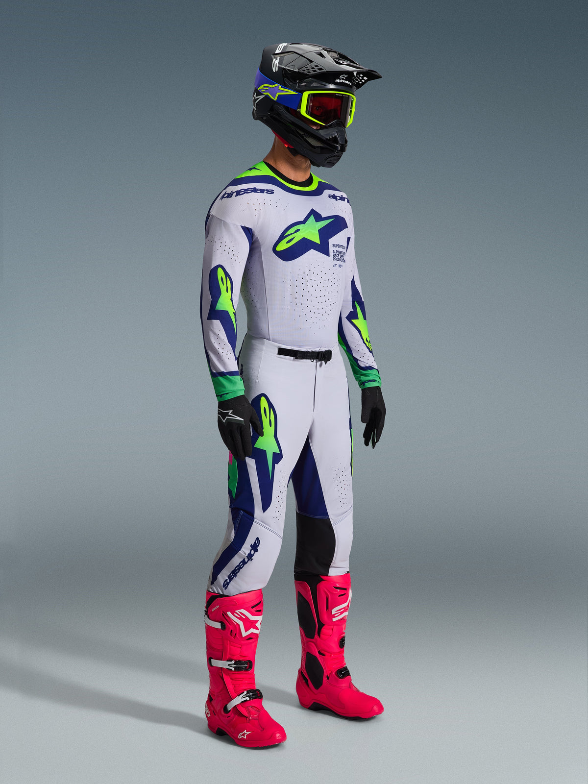 Alpinestars 2026 Supertech Vista Gear Set, Motocross rider in complete racing outfit, white jersey and pants with lime green and navy blue accents, bright pink boots, black helmet with blue and lime details, coordinated pro-level off-road apparel