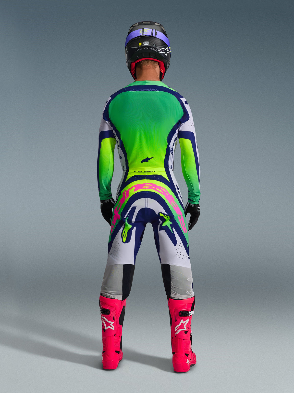 Alpinestars 2026 Supertech Vista Gear Set, Motocross gear set rear view, green and yellow gradient jersey, white and blue pants with a large pink logo on the back, bright pink boots and a purple helmet, professional off-road racing apparel