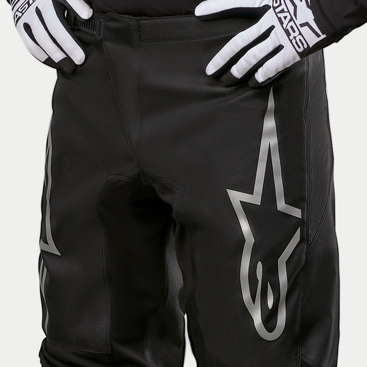 Alpinestars 2024 Fluid Graphite Pants, Motocross Pants, Black and Silver, detail shot of the upper thighs and waist, featuring a large silver star logo on the leg, worn with black and white racing gloves