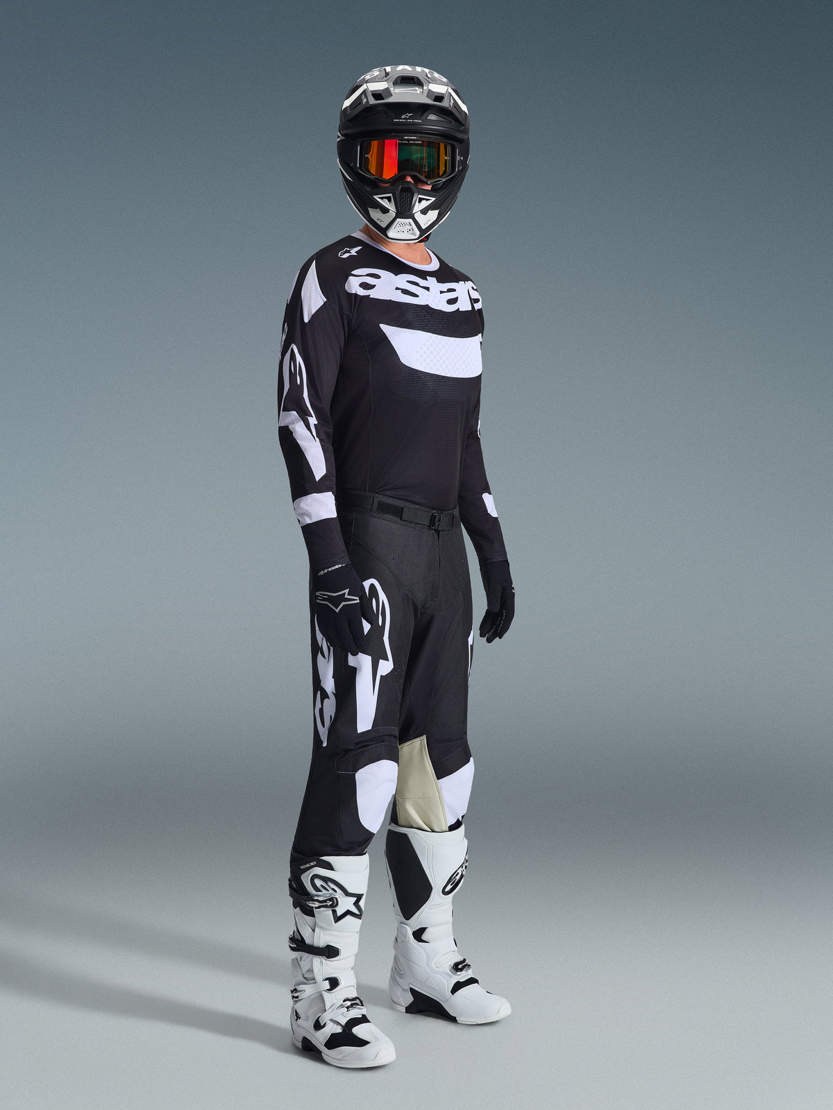 Motocross rider wearing complete racing gear, 2026 Racer Air Riway Jersey in Black and White, matching black pants with large white Alpinestars logos, black helmet with white accents and orange goggles, white and black boots, coordinated outfit for professional off-road racing