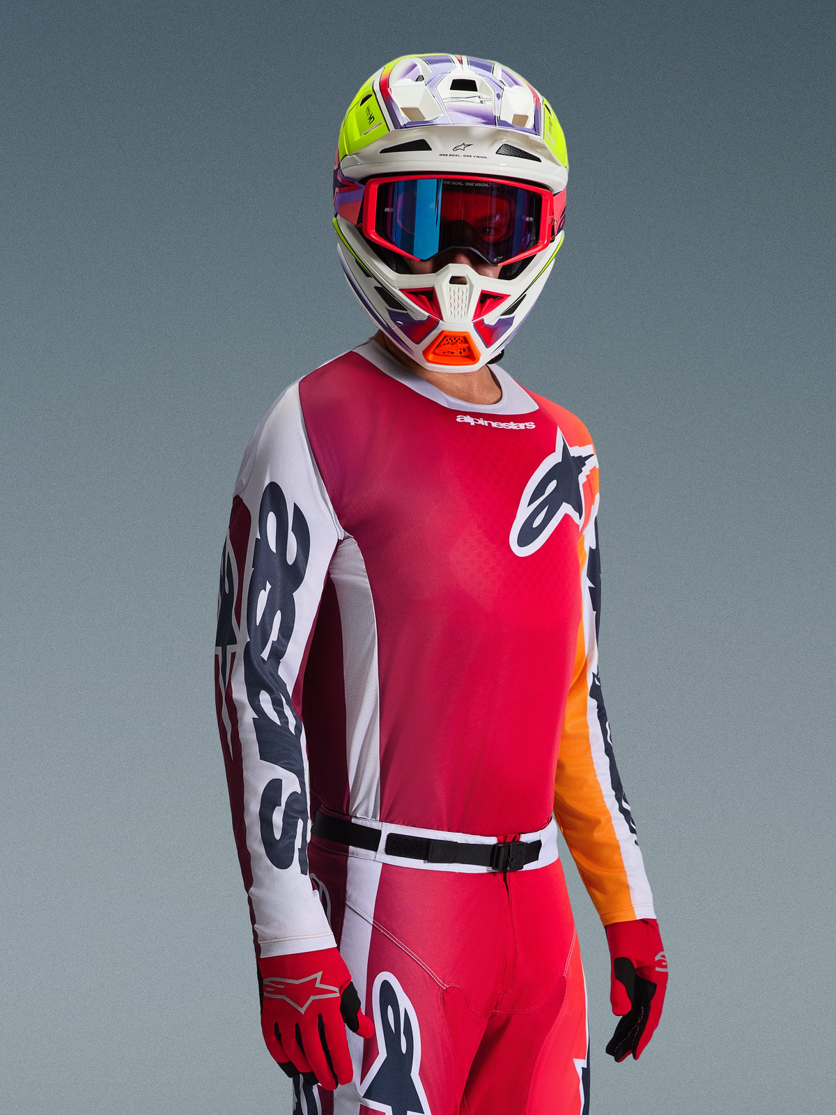 Motocross rider wearing the Alpinestars 2026 Racer Portl Gear Set, featuring a red jersey with pink tones and white sleeves, matching red pants, white helmet with vibrant yellow and purple accents, red goggles with blue iridescent lenses, and red gloves, professional racing outfit for motocross.