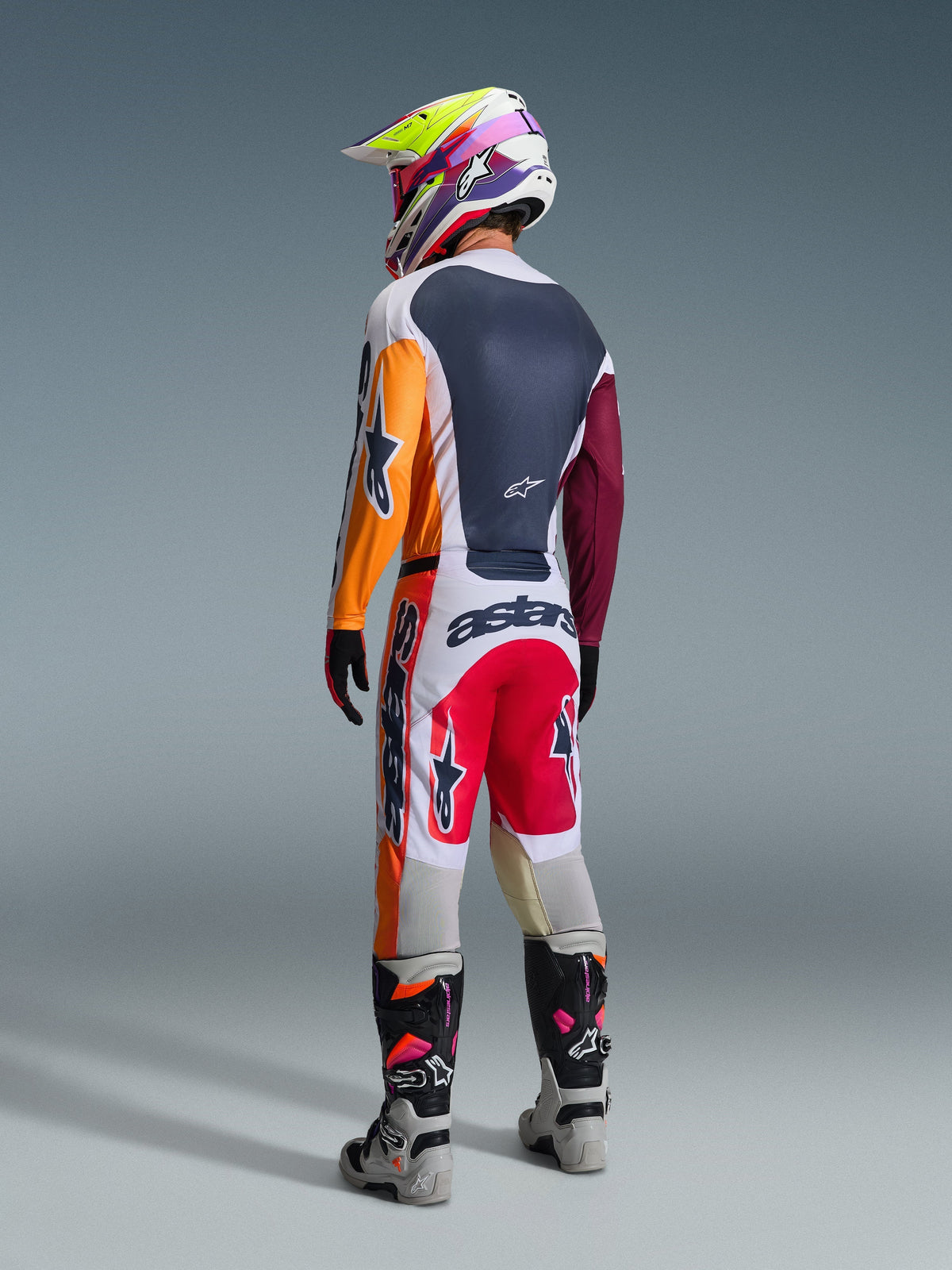 Motocross rider from behind wearing the Alpinestars 2026 Racer Portl Gear Set, featuring a color-blocked jersey with orange and maroon sleeves, white pants with red panels and bold 'astars' branding, multi-colored helmet, and gray boots, professional off-road racing outfit.