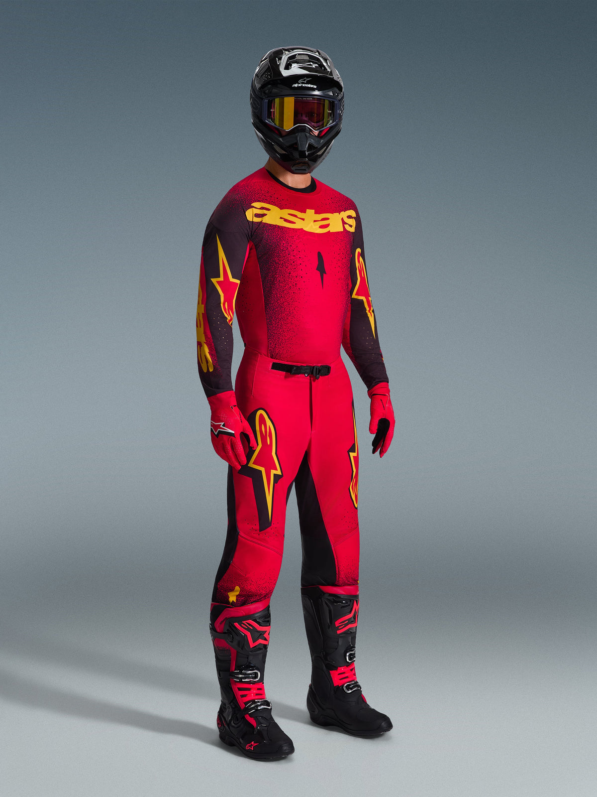 Motocross rider wearing complete racing gear, red jersey with black sleeves and yellow graphics, matching red pants with black panels and yellow logos, black helmet with yellow-lens goggles, black and red boots, red and black gloves, coordinated 2026 Supertech Scenz outfit for professional motocross racing