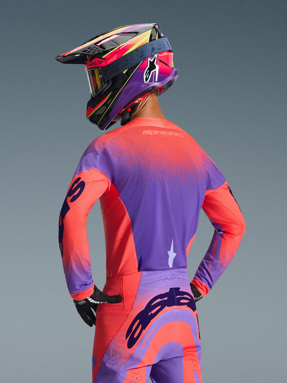 Motocross rider from behind wearing the 2026 Alpinestars Supertech Scenz Jersey in Hot Coral and Purple, featuring a vibrant speckled gradient design, paired with matching motocross pants and a colorful racing helmet, professional gear for off-road performance.