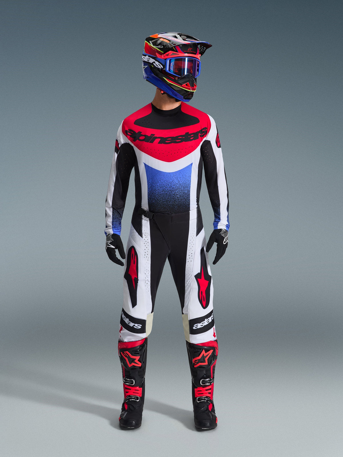 Motocross rider wearing the Alpinestars 2026 Techstar Knif Gear Set, black white and red jersey with blue gradient and branding, matching black and white pants with red accents, multi-colored helmet with blue goggles, and black and red boots, coordinated off-road racing outfit.