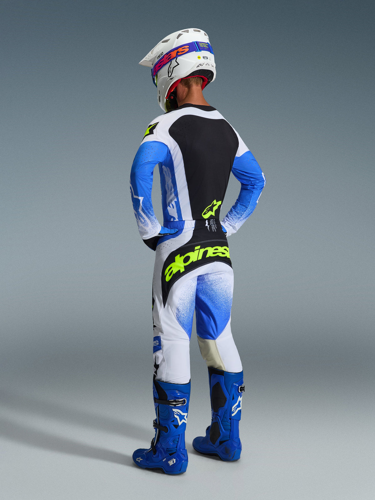 Motocross rider viewed from behind wearing the complete Alpinestars 2026 Techstar Knif Gear Set in Ucla Blue and Black, featuring a jersey with a black back panel and yellow fluo logo, matching blue and white pants with bold yellow branding, blue motocross boots, and a white racing helmet.