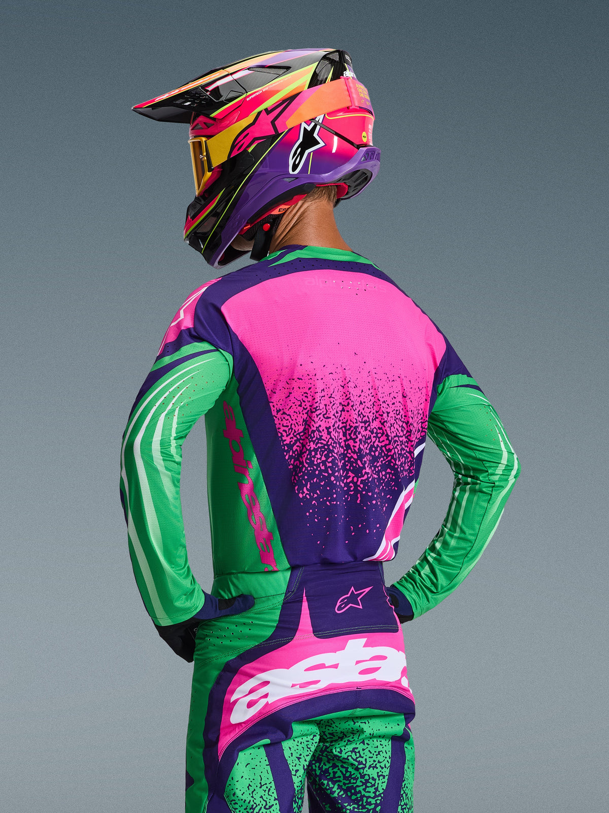 Motocross rider seen from the back wearing the Alpinestars 2026 Techstar Nomur Gear Set, Deep Purple Green and Pink Fluo jersey and matching pants, featuring bold graphics and an athletic race fit, paired with a multi-colored helmet and yellow goggles.